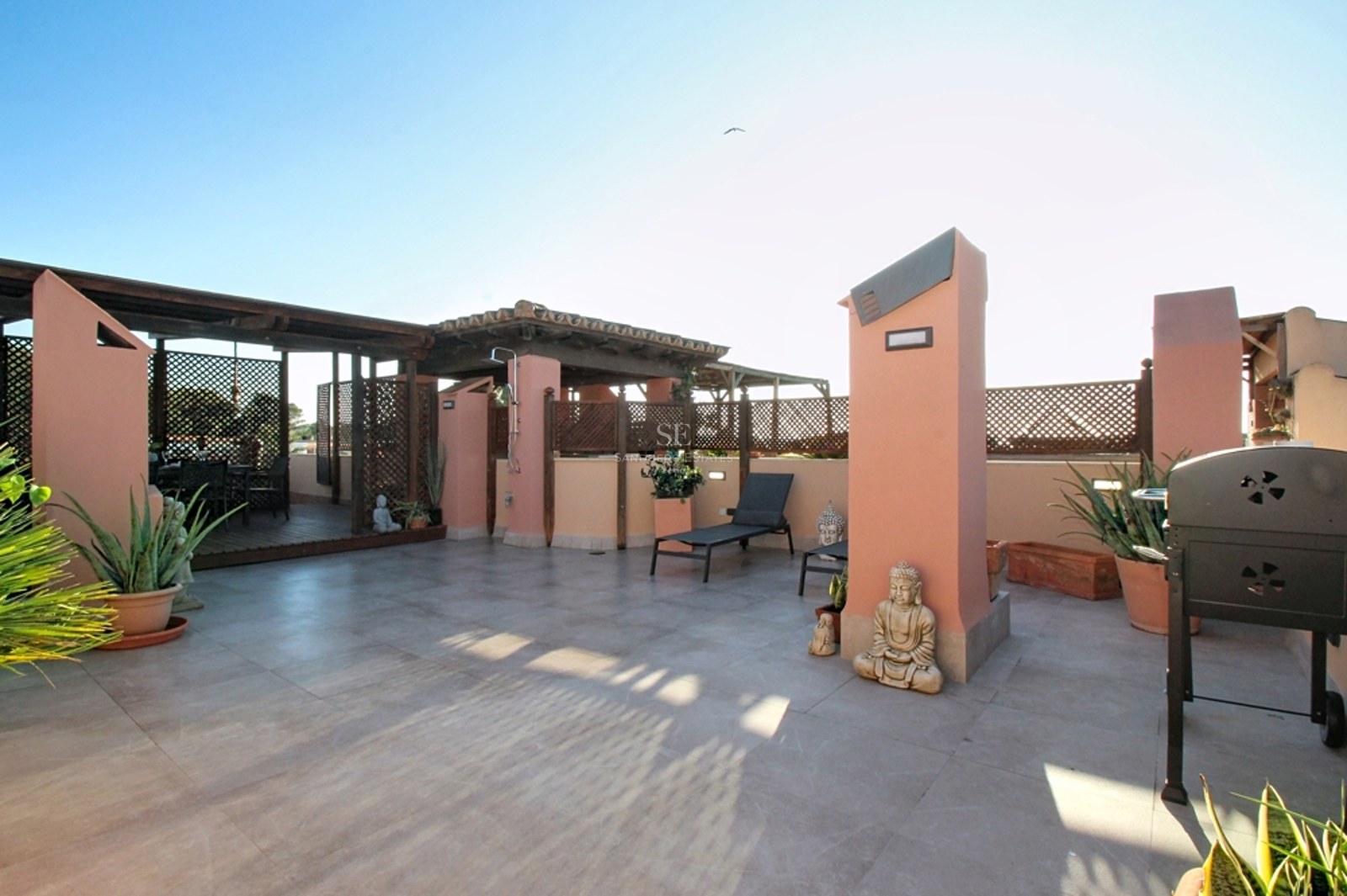 Terraza mediterránea en azotea con paredes terracota, pérgola de madera, suelo de piedra y tumbonas bajo un cielo despejado.