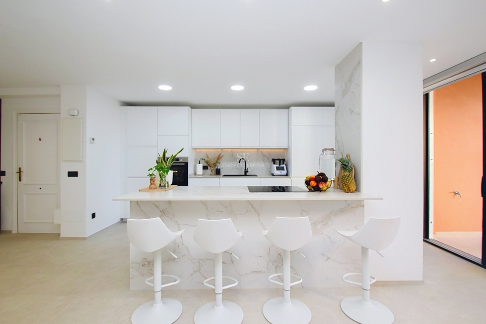 Cocina blanca moderna con una gran isla de mármol, cuatro taburetes blancos y electrodomésticos integrados.