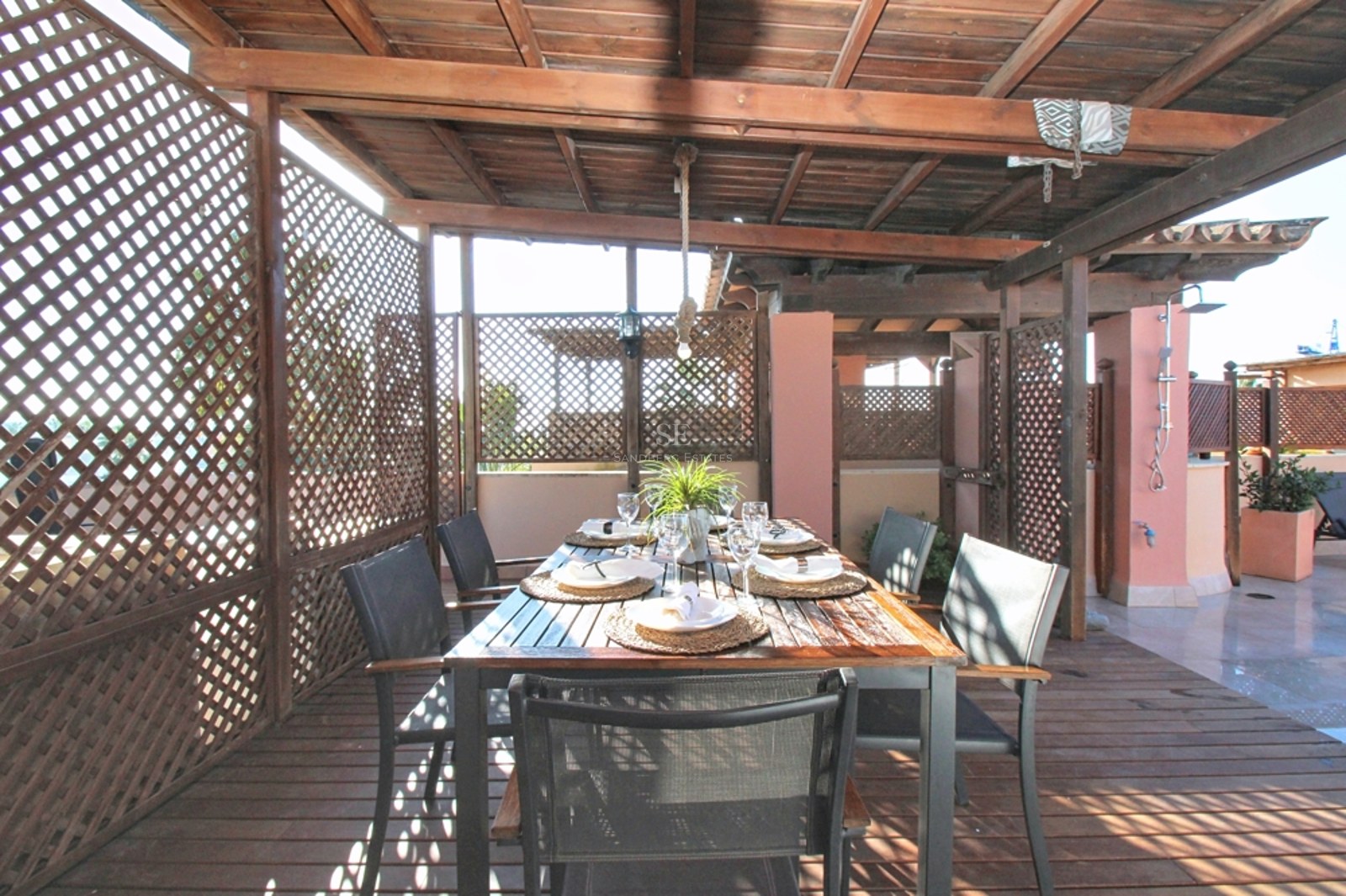 Mesa de comedor bajo pérgola de madera con celosías de privacidad y suelos de tarima en una terraza soleada.