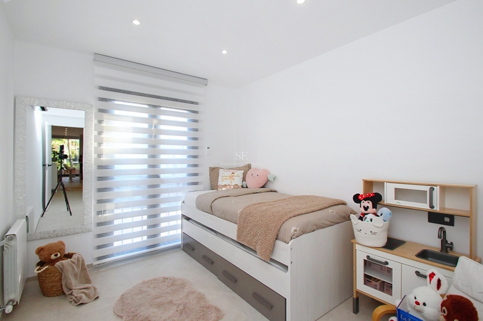 Dormitorio blanco con cama nido, espejo de cuerpo entero, persianas tipo cebra y zona de juegos infantil.