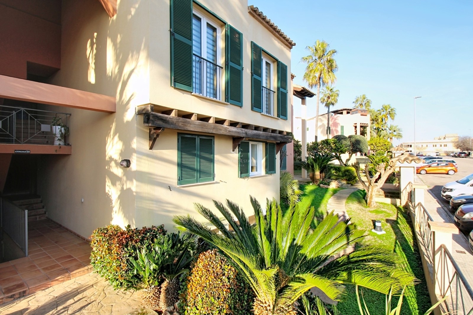 Exterior de edificio beige con persianas verdes, vigas de madera y jardín tropical con palmeras.