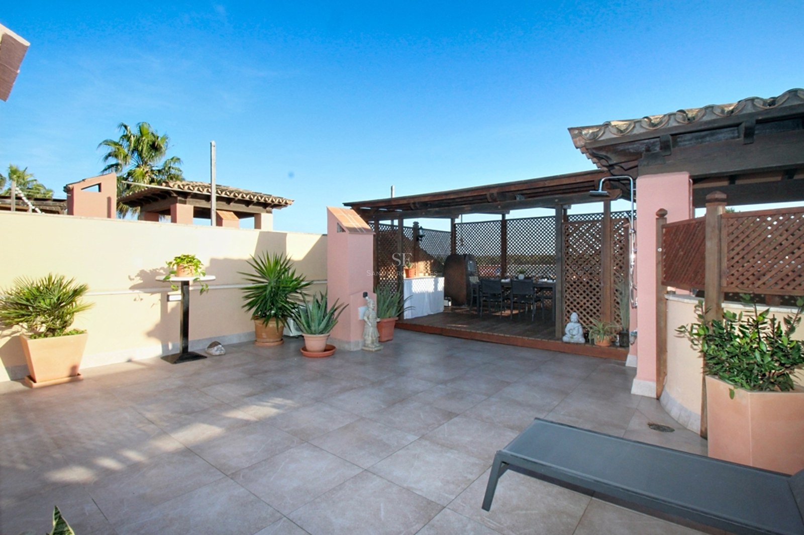 Terraza soleada con suelo de baldosas grises, pérgola de madera, paredes terracota y plantas ornamentales.