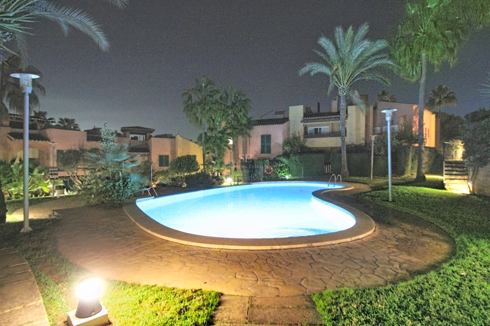 Piscina con forma de riñón iluminada de noche, rodeada de palmeras, césped y villas mediterráneas bajo un cielo oscuro.