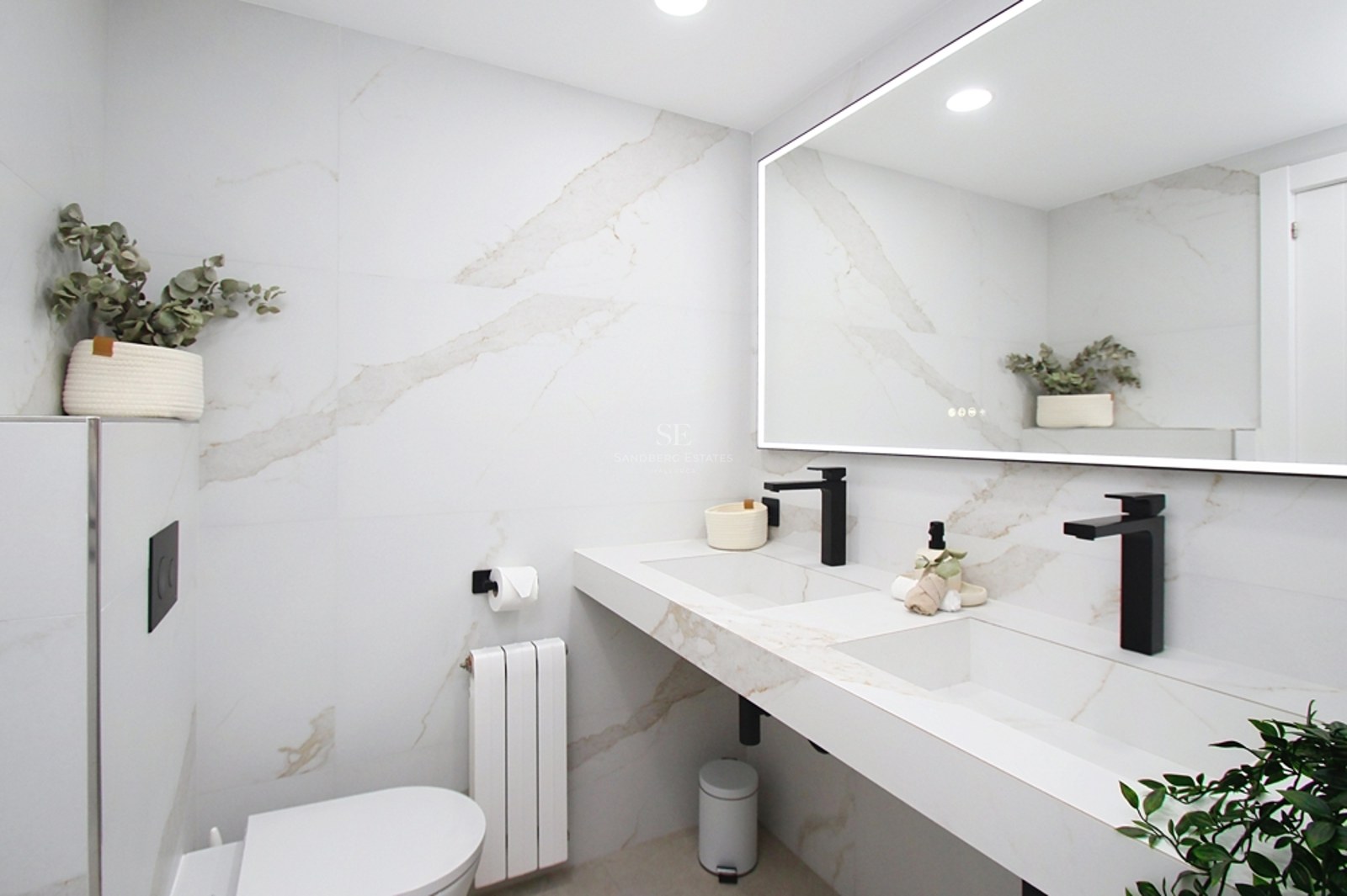 Baño moderno con paredes efecto mármol blanco, lavabo doble con grifería negra mate y gran espejo retroiluminado.