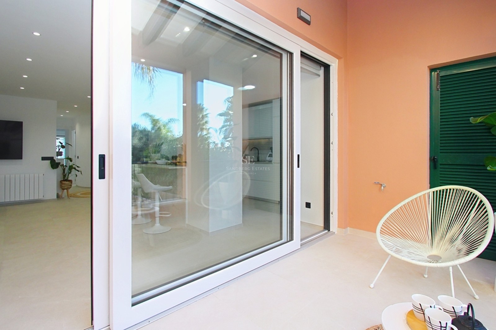 Terraza luminosa con silla Acapulco blanca y puertas correderas de cristal que abren hacia un salón moderno.