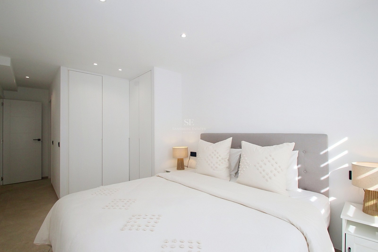 Un dormitorio moderno y luminoso con ropa de cama blanca, cabecero gris, armarios integrados y luz natural.