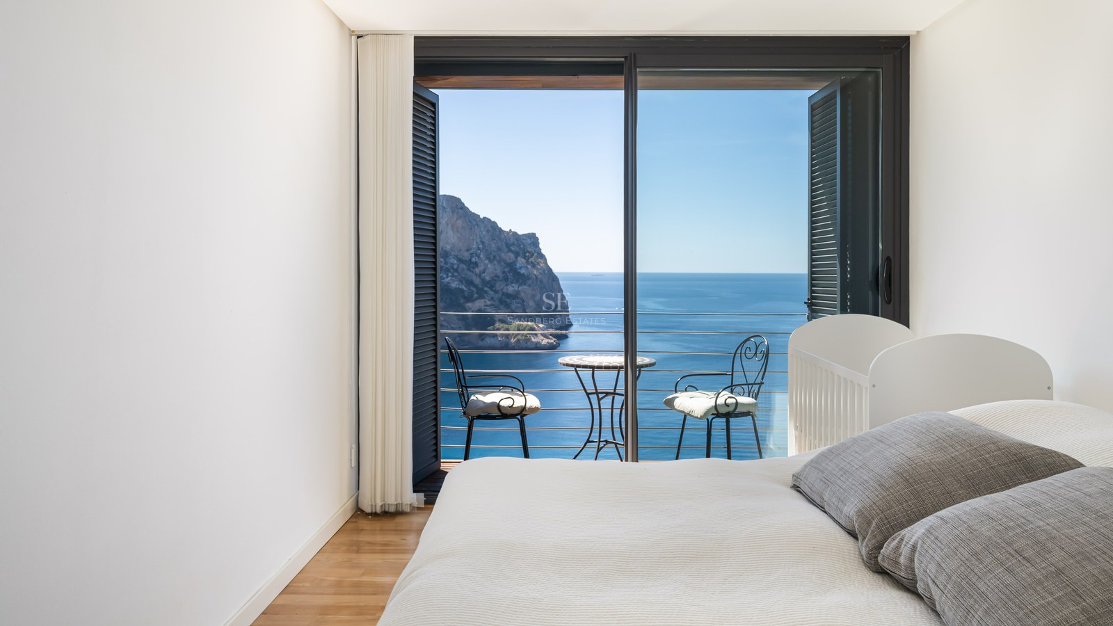 Chambre minimaliste blanche avec balcon privé, mobilier en fer forgé et vue sur une falaise et la mer Méditerranée.