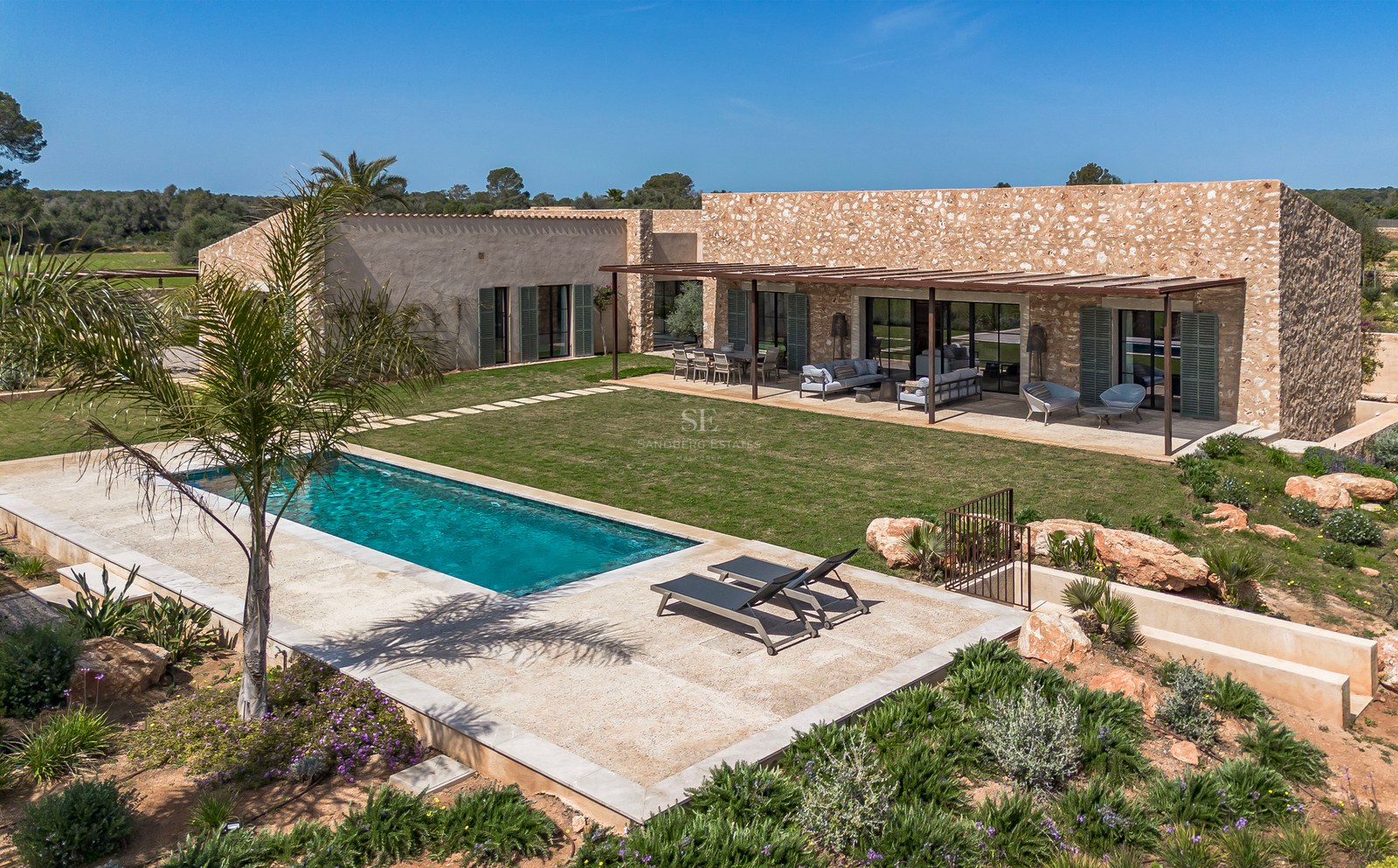 Außenansicht einer Steinvilla mit rechteckigem Pool, Rasenfläche und Pergola-Terrasse in Mallorca.
