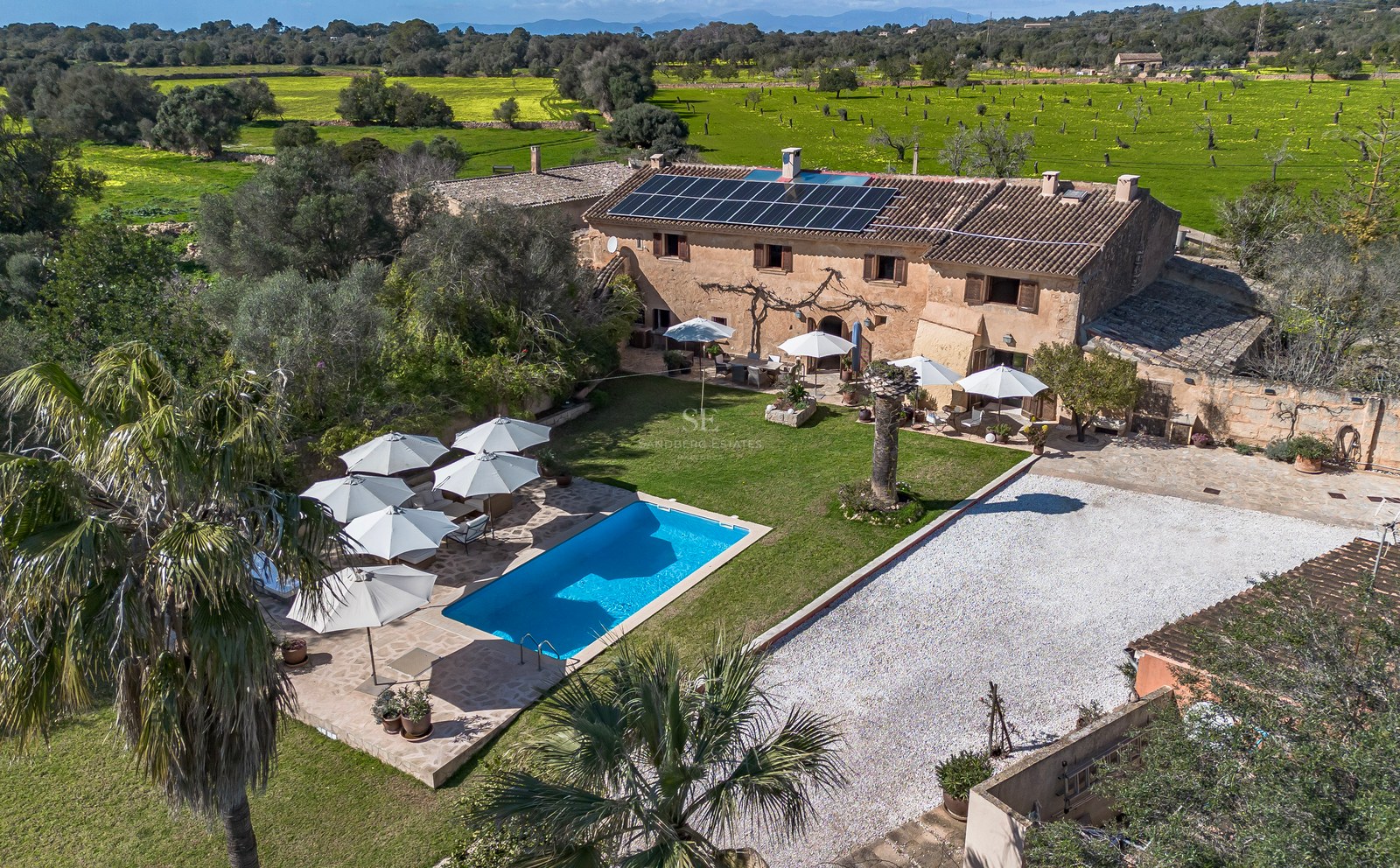 Luftaufnahme einer Steinvilla mit blauem Pool, Sonnenkollektoren und weißen Sonnenschirmen, umgeben von Feldern.