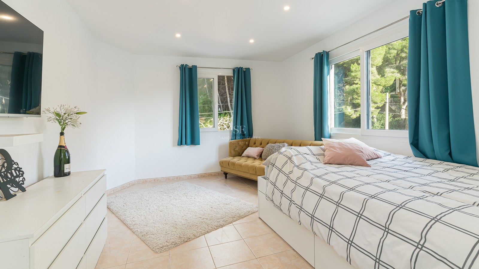 Bright bedroom with a bed, mustard yellow velvet sofa, teal curtains, and large windows looking out to trees.