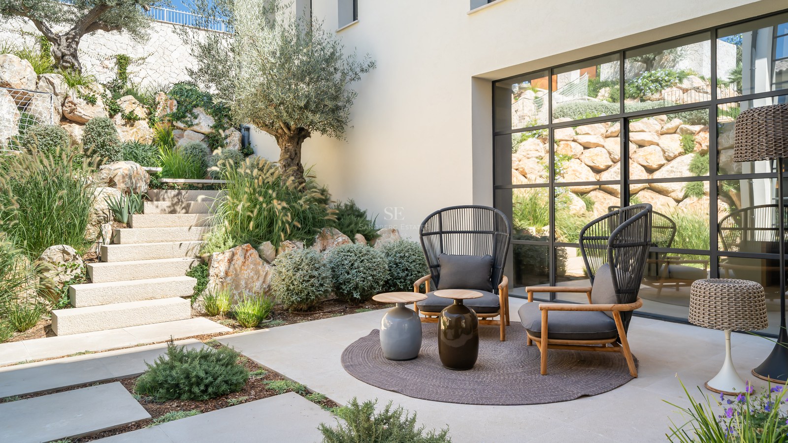 Modern patio with two grey armchairs, stone steps, olive tree, and large industrial-style window.