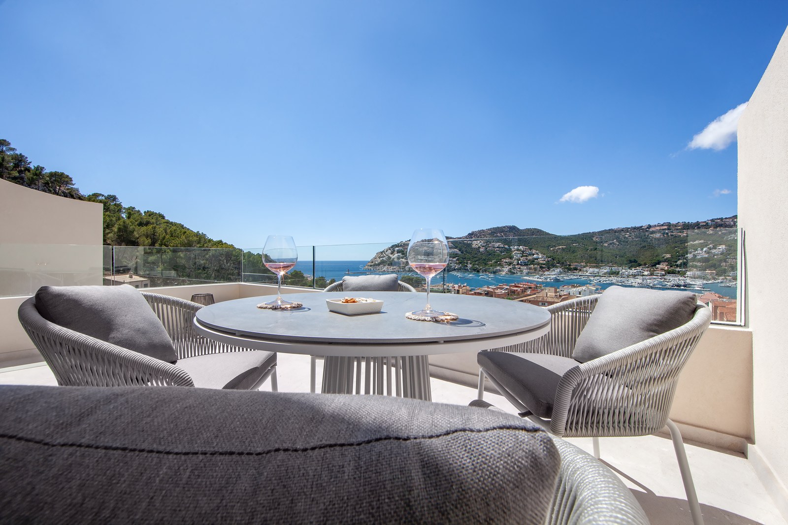 Modern terrace table with two glasses of wine set against a spectacular view of the harbor and sea under a clear blue sky.