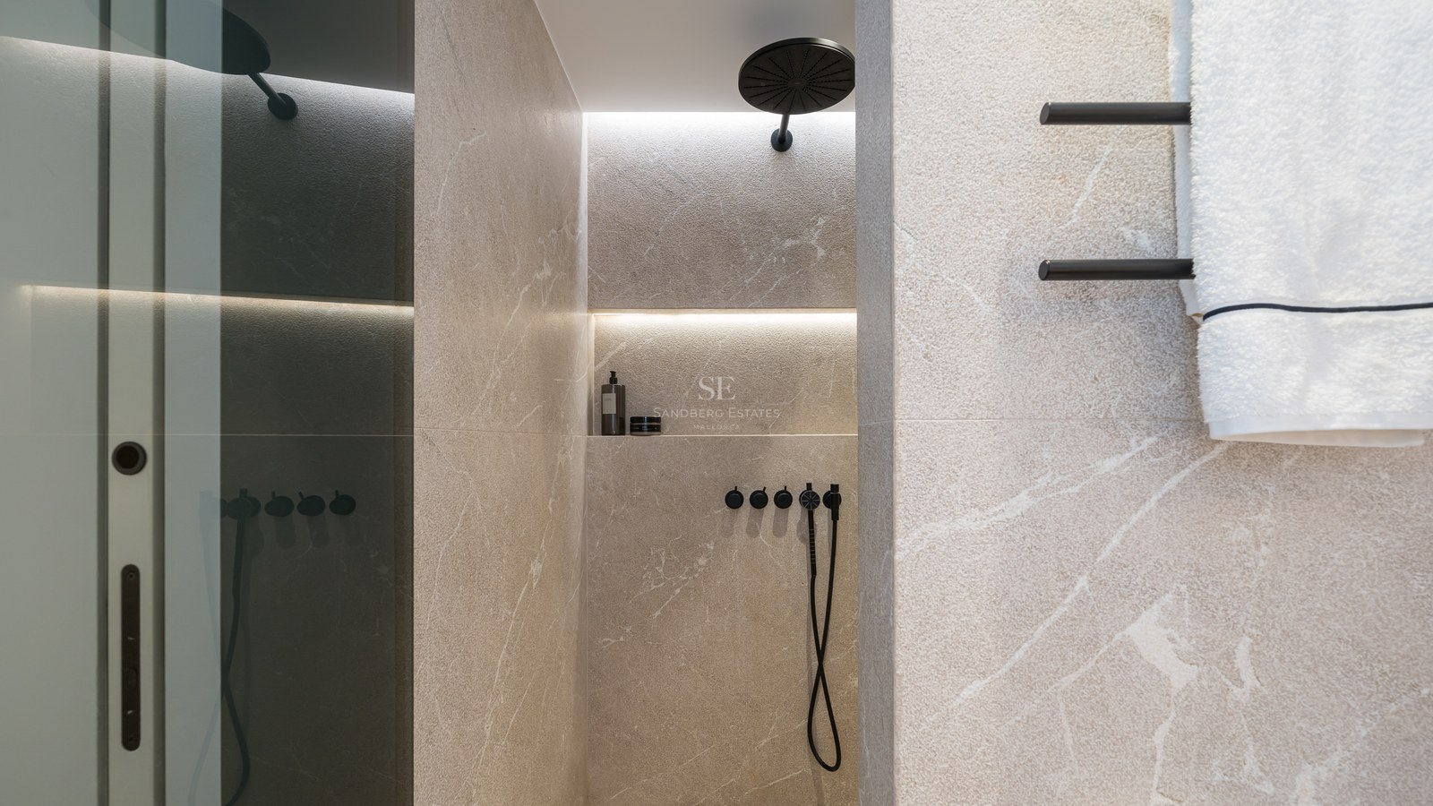 Douche avec carrelage en pierre beige, pommeau de pluie noir mat et niche éclairée sur un mur en pierre veinée.
