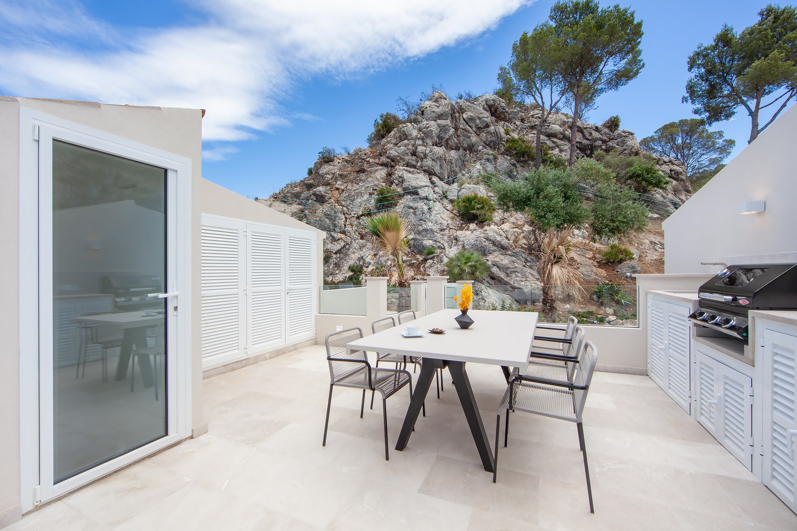 A modern white terrace with a dining table, chairs, and a barbecue area, set against a rocky mountain backdrop.