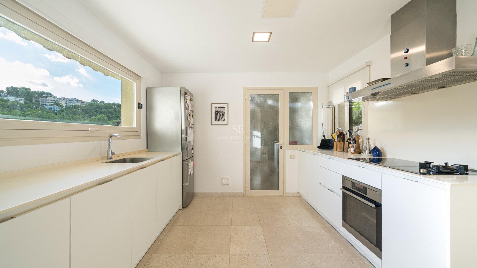 Bright kitchen with white cabinets, stainless steel fridge and oven, and a large window overlooking green trees.