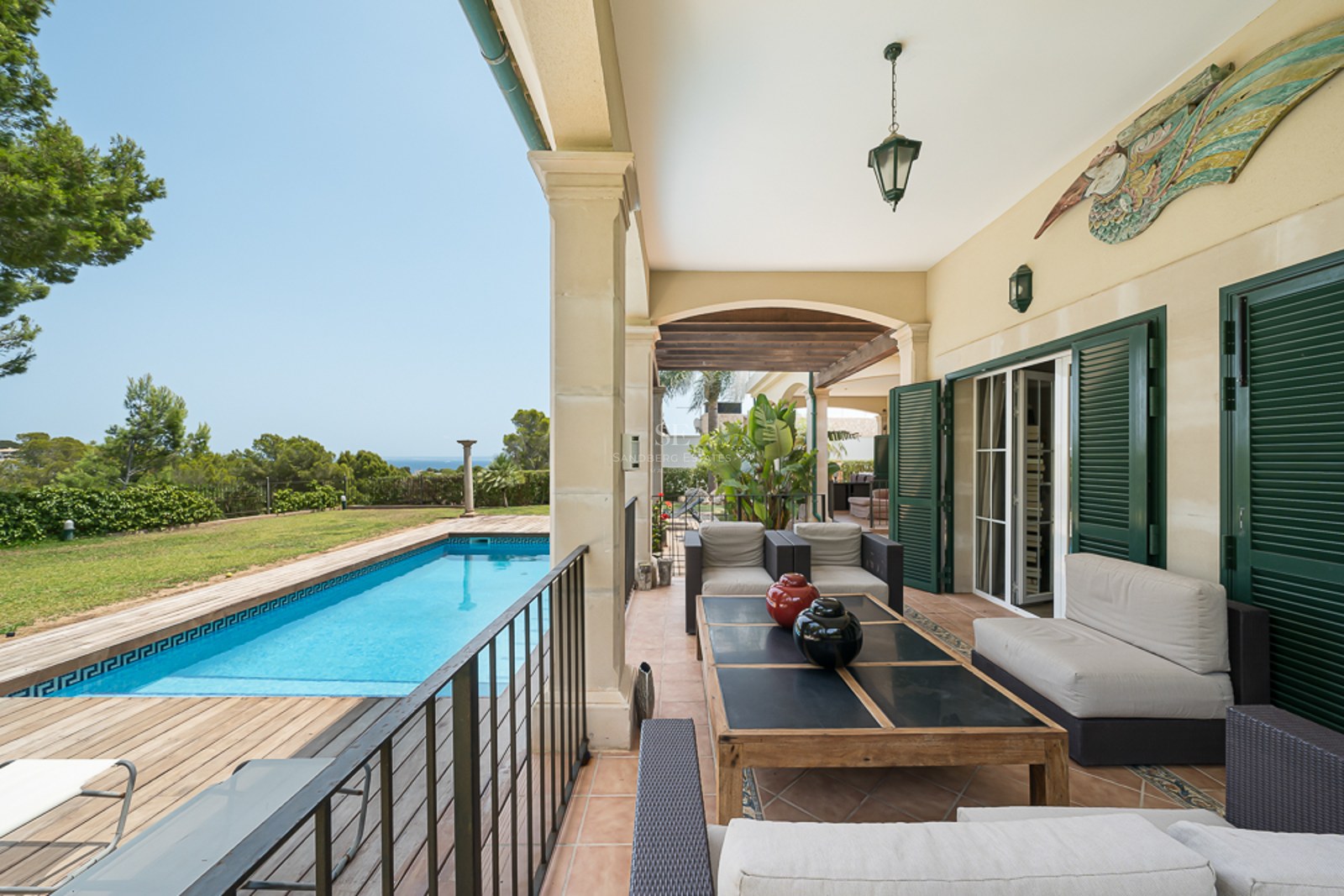 Porche couvert avec salon de jardin donnant sur une piscine, une terrasse en bois et la mer au loin.