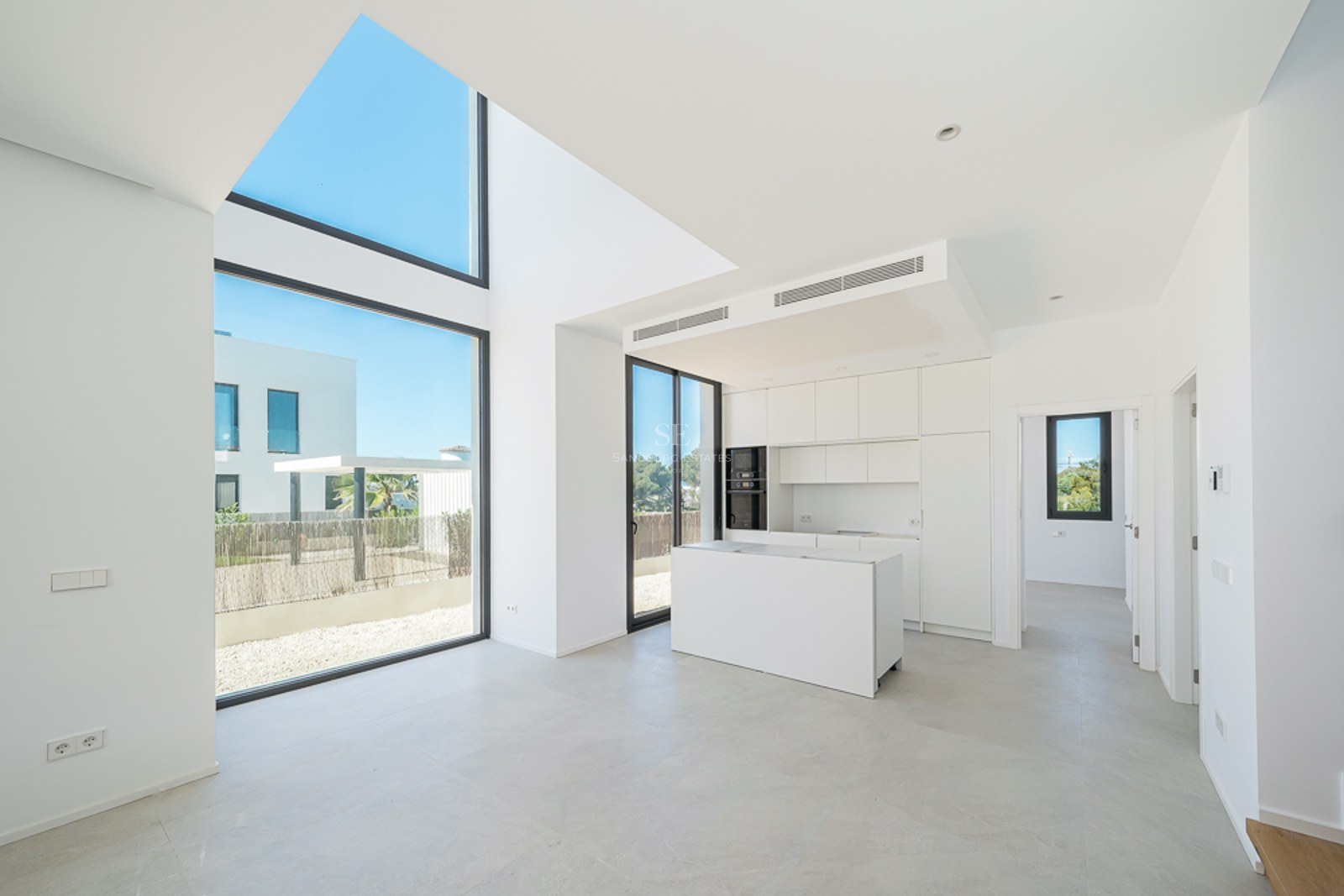 Cuisine blanche minimaliste avec îlot, baies vitrées double hauteur et lumière naturelle abondante.