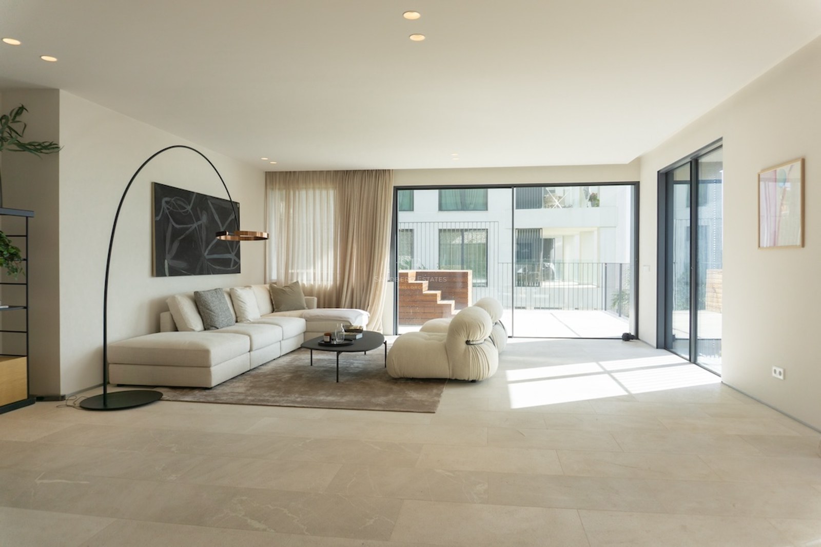 Ljust minimalistiskt vardagsrum med beige stengolv, off-white hörnsoffa och stora skjutdörrar i glas.