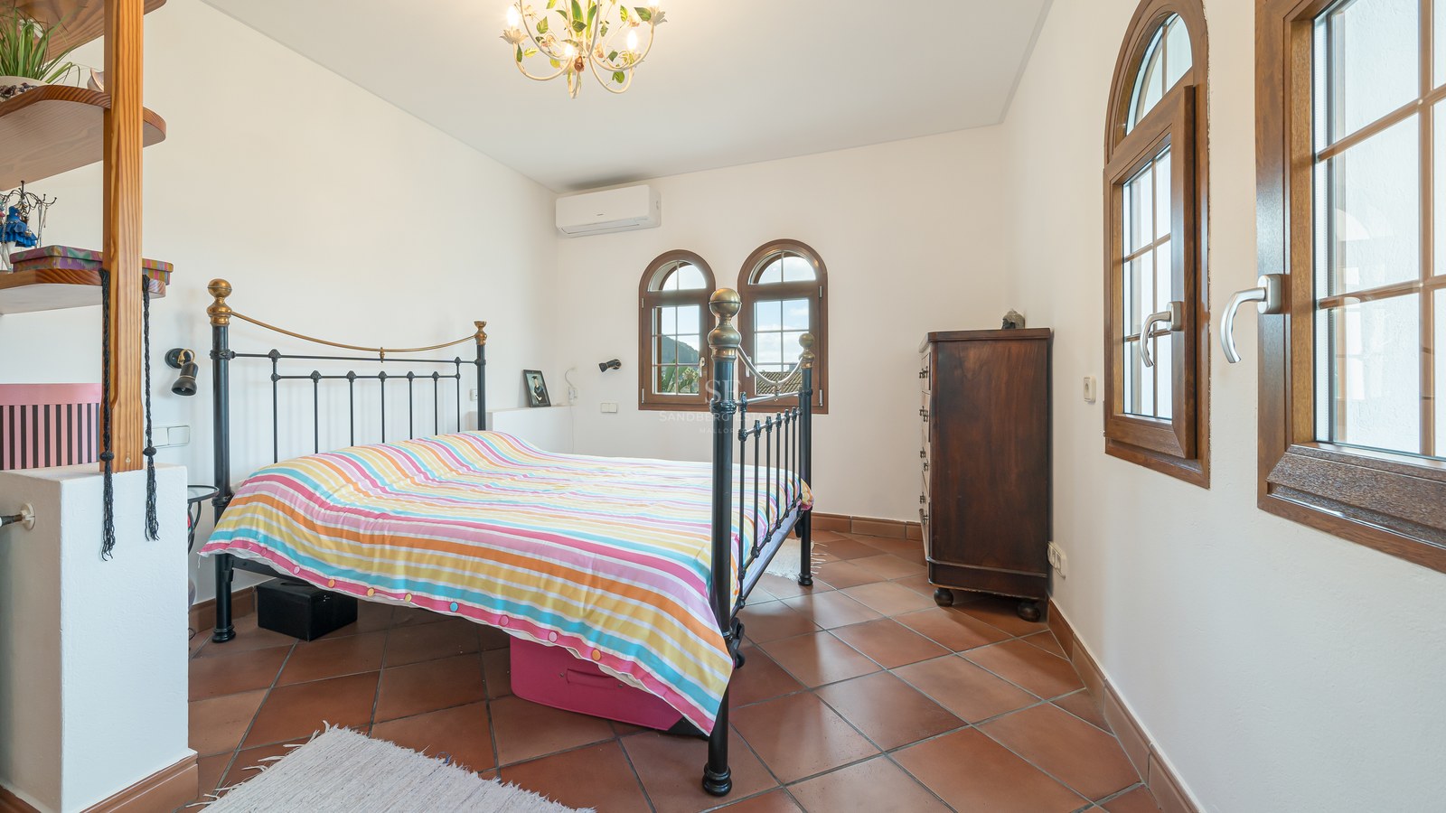 Chambre lumineuse avec sol en terre cuite, lit en fer forgé et fenêtres traditionnelles en bois en forme d'arche.