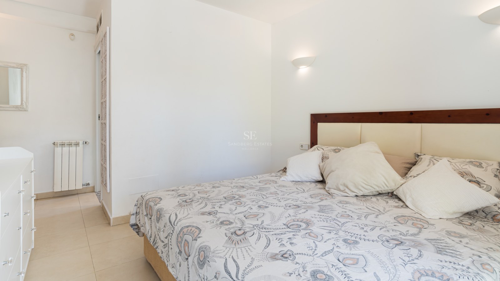 Dormitorio con sábanas estampadas, paredes blancas, suelo de gres crema y cabecero de madera con apliques.