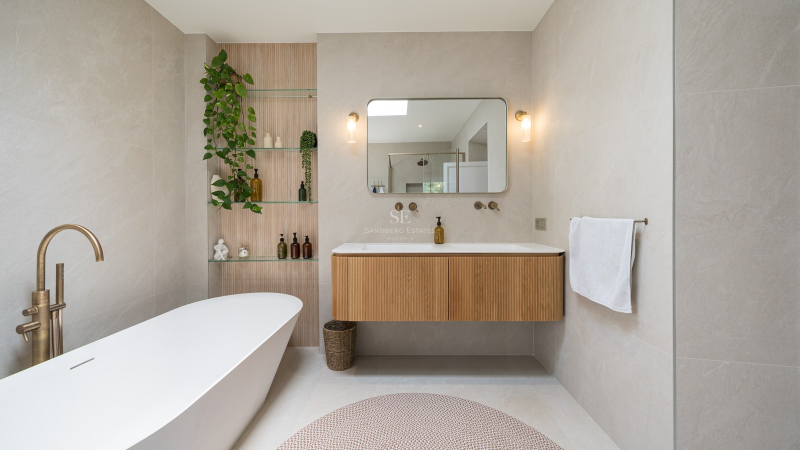 Contemporary bathroom featuring a white freestanding bathtub, floating fluted wood vanity, and brass fixtures against beige stone walls.