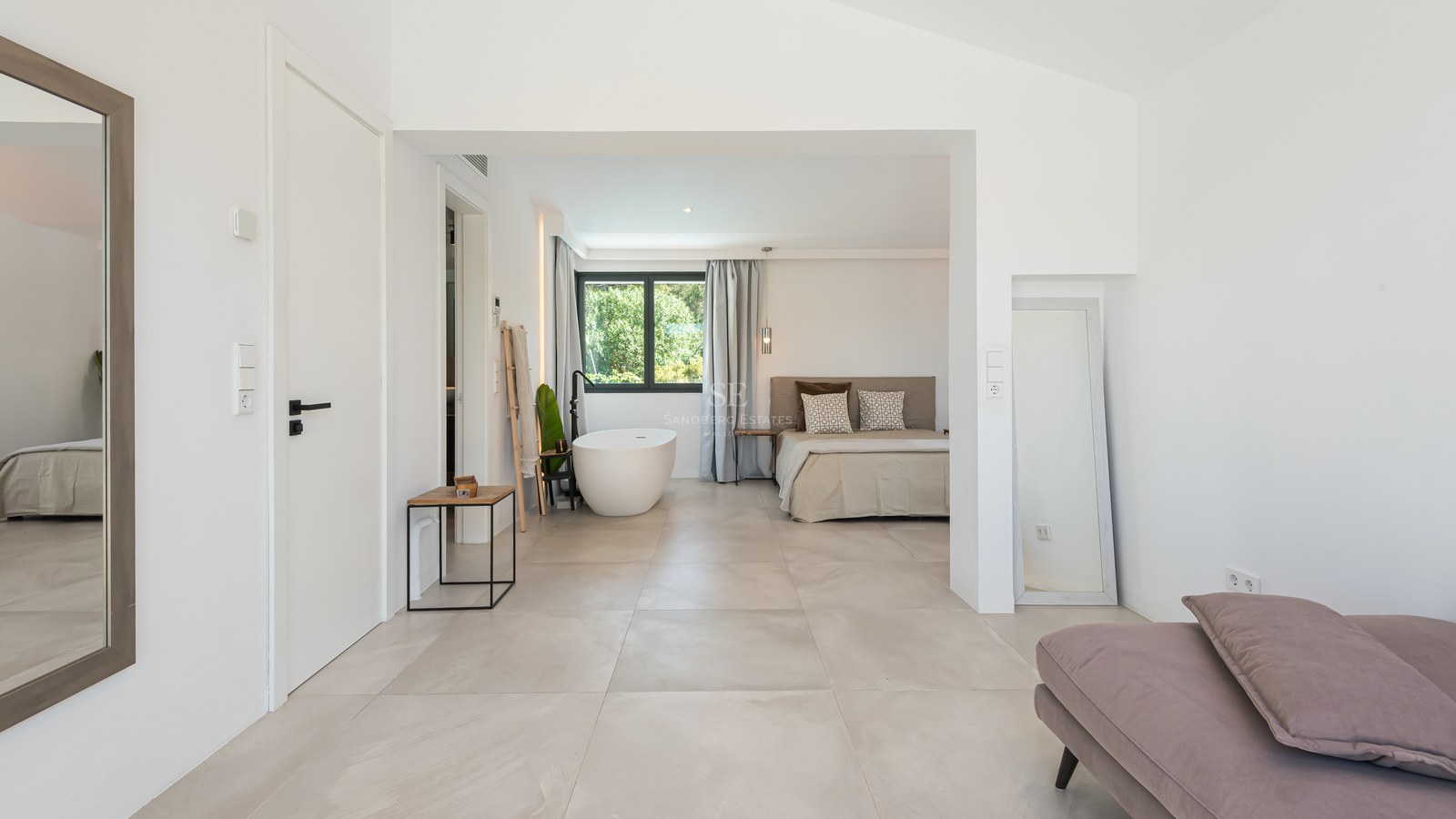 A bright master bedroom featuring large format floor tiles, a minimalist bed, and a white freestanding bathtub by a window.