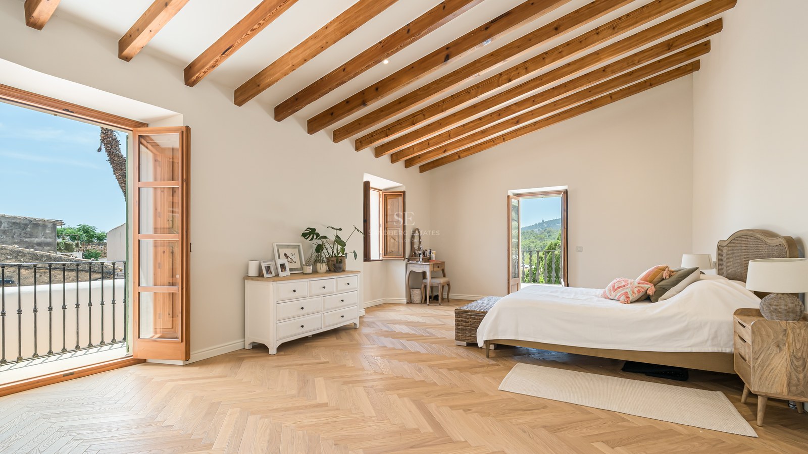 Spacious bedroom with high vaulted ceilings, wooden beams, herringbone oak floors, and a balcony overlooking a village.