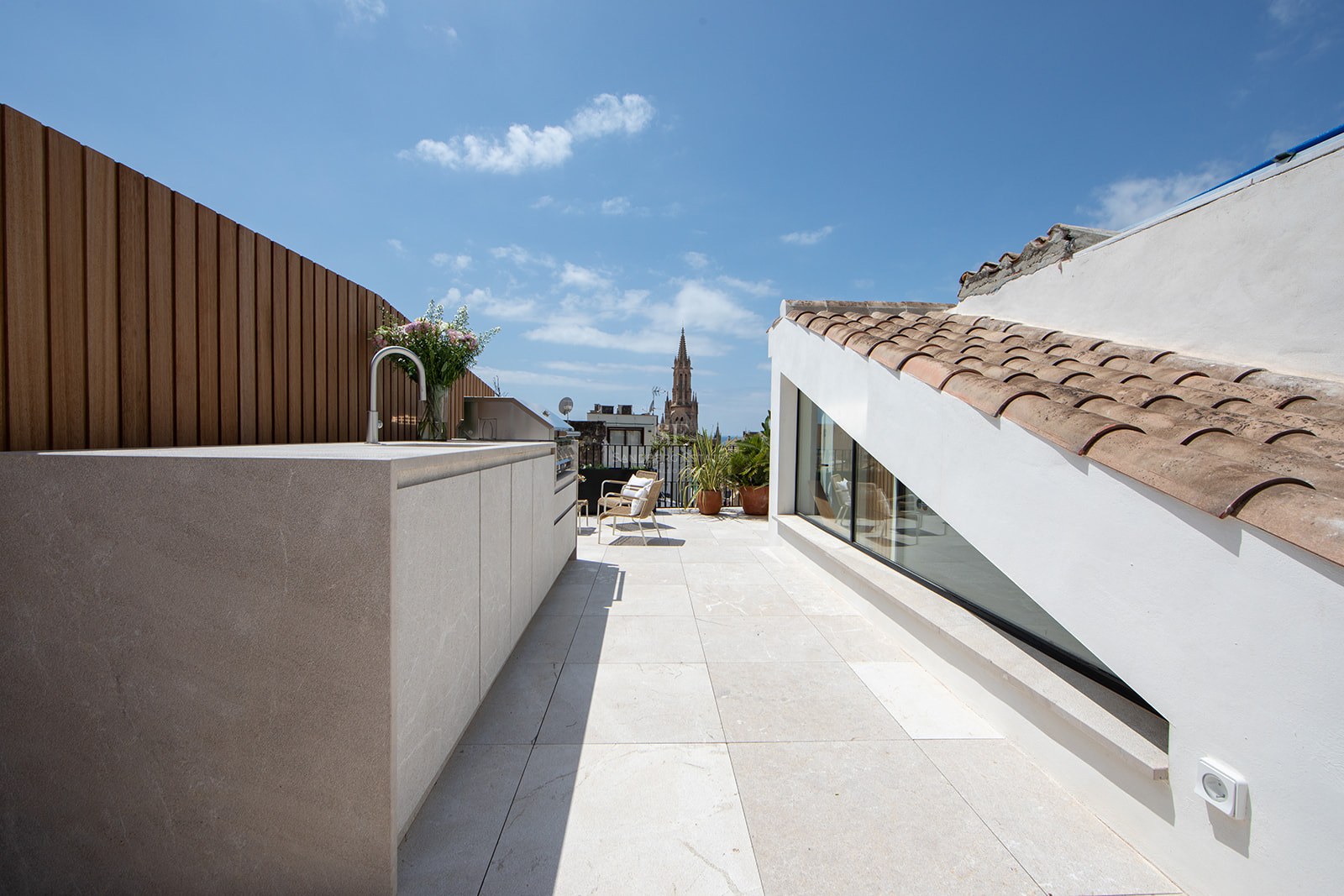 Modern rooftop terrace with outdoor kitchen, stone flooring, and a cathedral spire view under a blue sky.