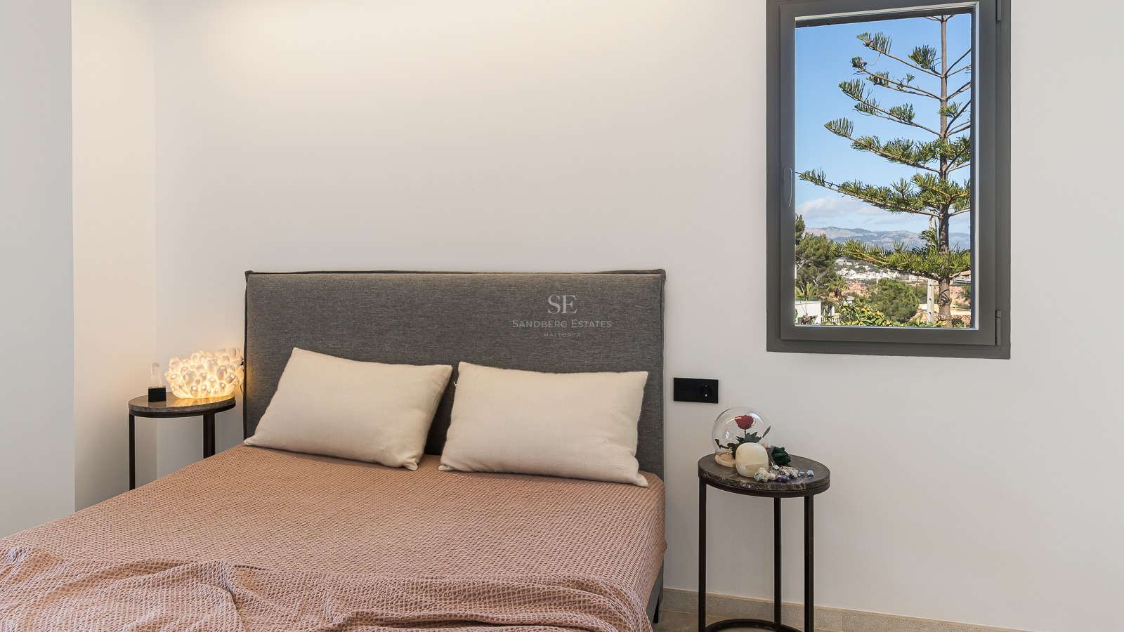 Bett mit grauem Kopfteil, terrakottafarbener Decke und Marmortischen vor Fenster mit Blick auf Berge und Pinien.