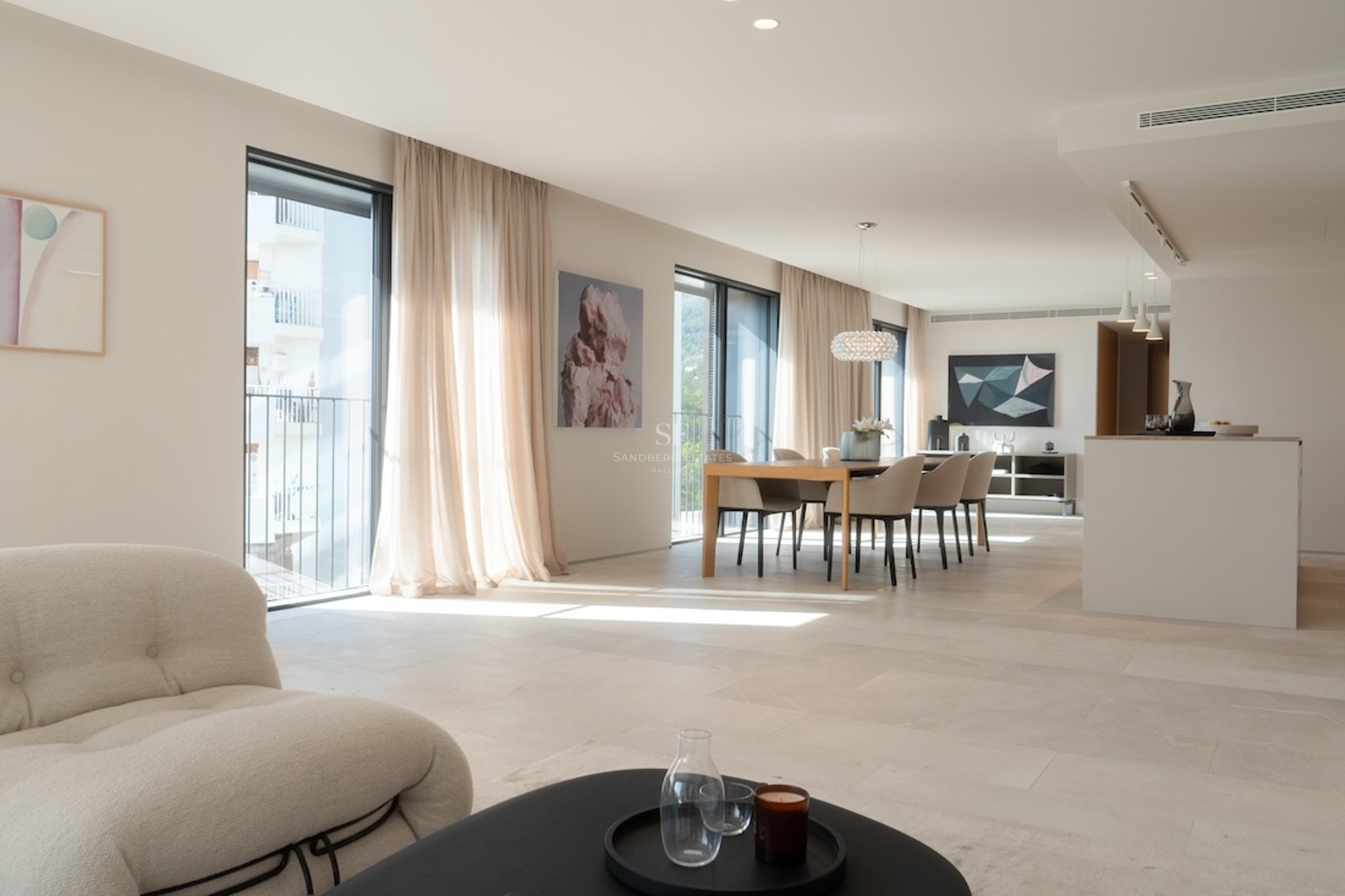 Ljust vardagsrum med öppen planlösning, naturstensgolv, stora fönsterpartier och moderna möbler.