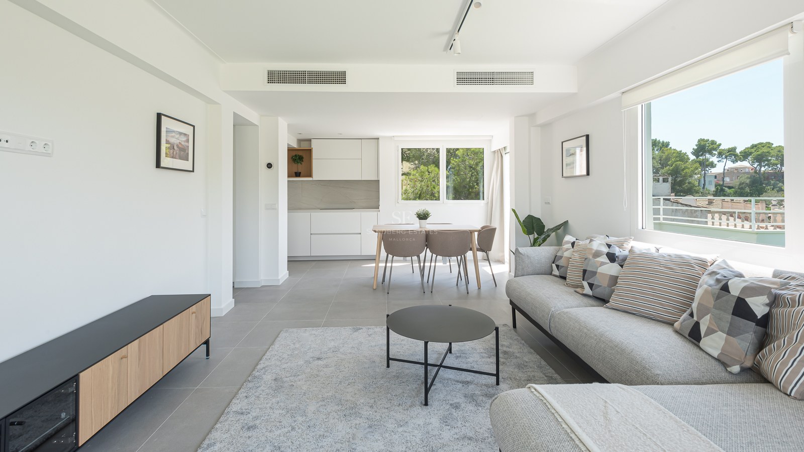Salon lumineux avec canapé gris, coin repas et cuisine blanche épurée avec vue sur la nature.