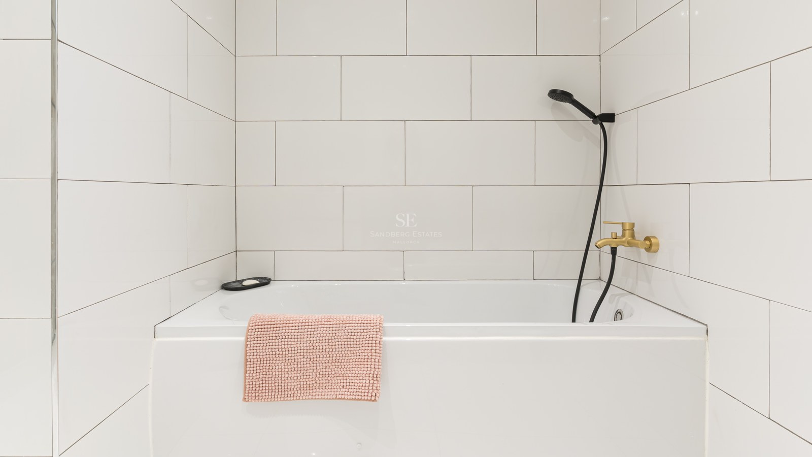 White bathtub with tiled walls, brass faucet, black shower head, and a textured pink bath mat.