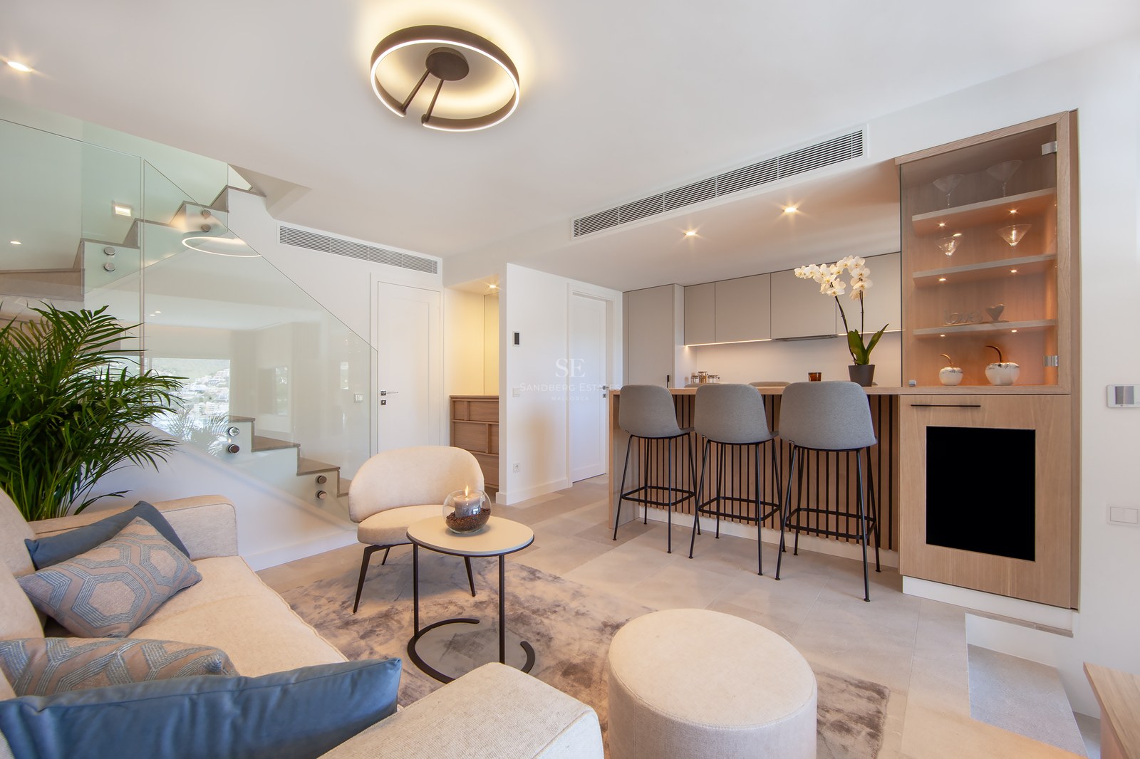 Modern living room with cream sofa, glass staircase, and a wood-paneled breakfast bar with grey stools.