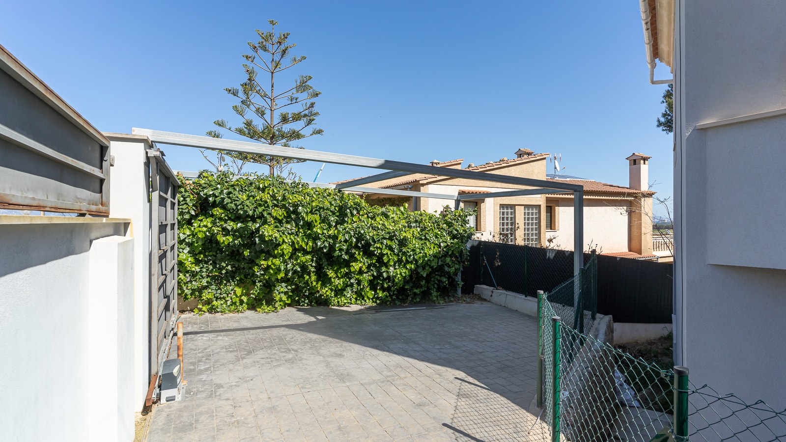 Gepflasterter Privatparkplatz mit automatischem Schiebetor und Metall-Carport-Rahmen vor blauem Himmel.