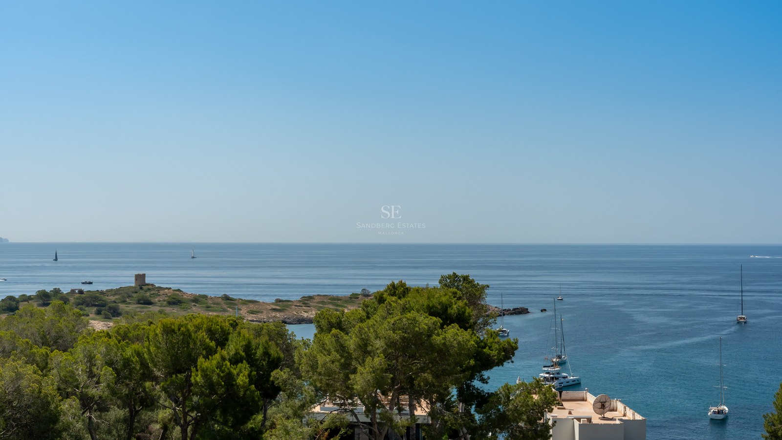 Wide view of a blue bay with sailboats, pine trees in the foreground, and a historic stone tower on a distant point.