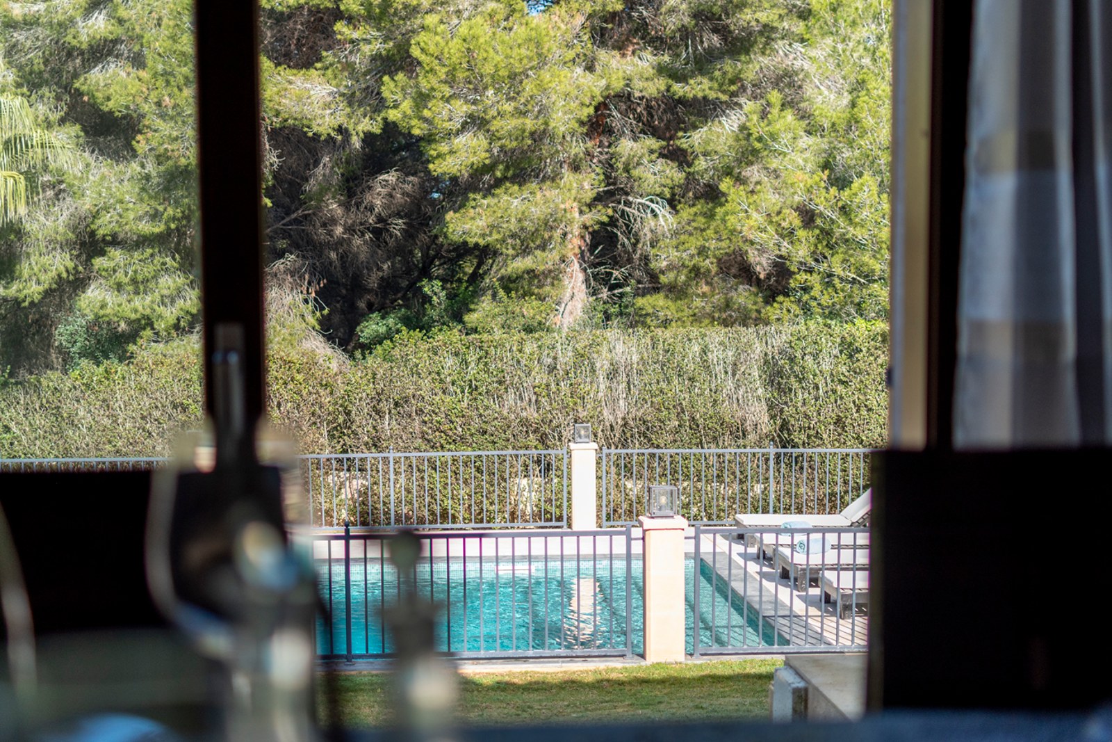 Vue par une fenêtre d'une piscine turquoise clôturée entourée de transats et de grands pins.