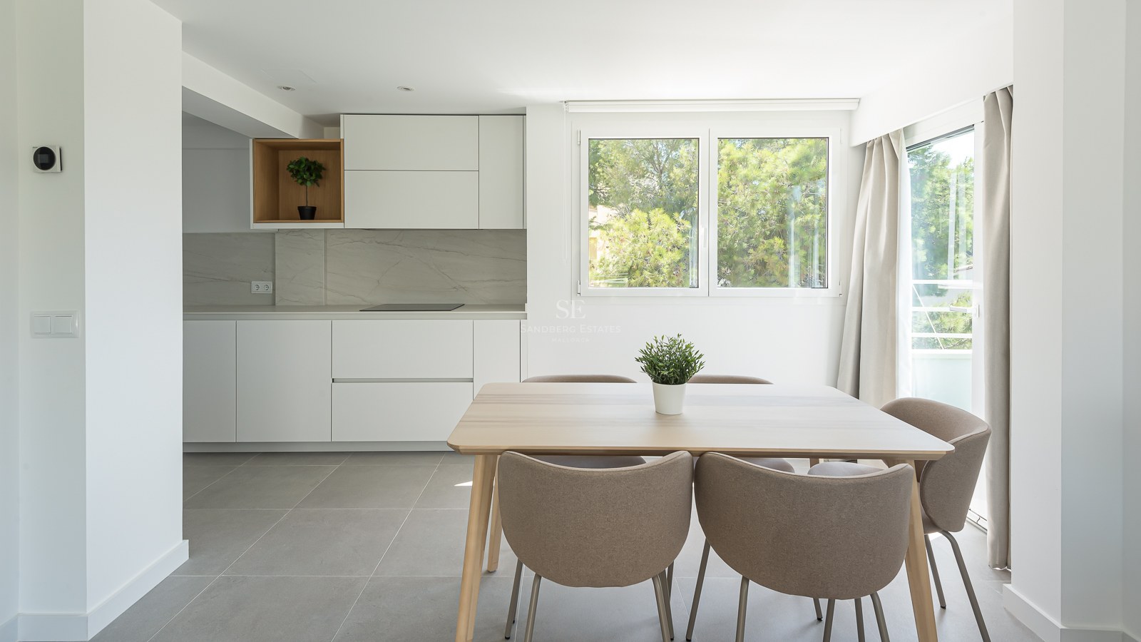 Salle à manger lumineuse avec table en bois clair, chaises taupe et cuisine blanche minimaliste vue sur jardin.