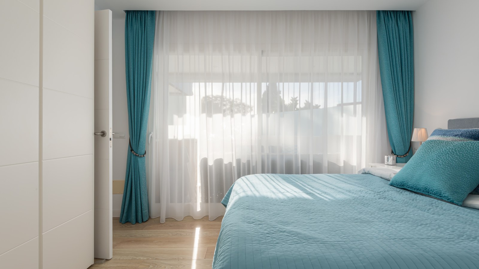 A bright bedroom with a teal bedspread and matching curtains, featuring floor-to-ceiling windows and wood flooring.