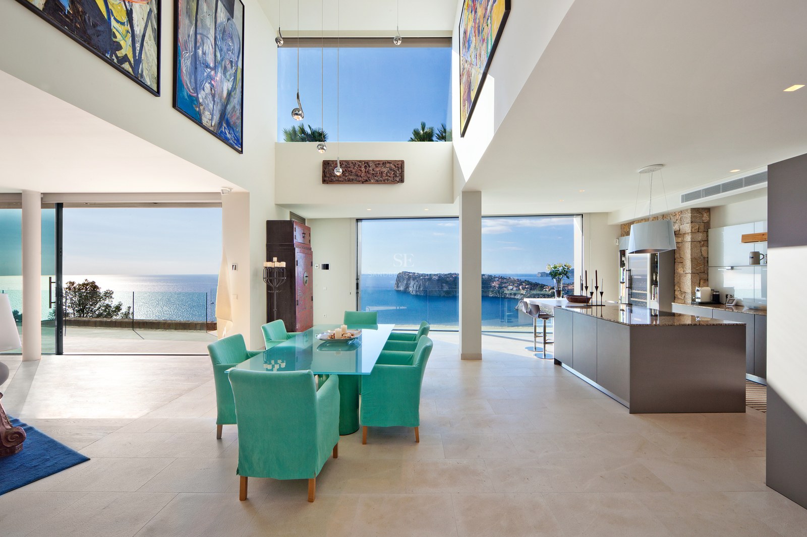 Modern open-plan dining and kitchen area with turquoise chairs and floor-to-ceiling windows overlooking the sea.