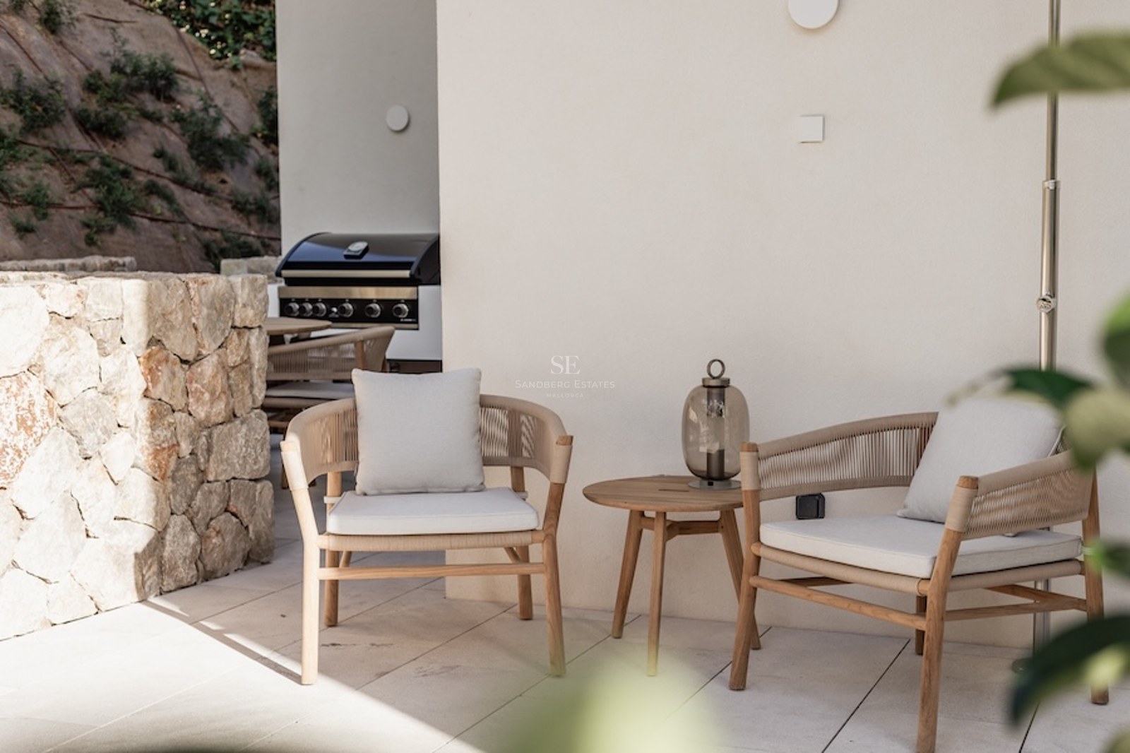 Deux élégants fauteuils modernes avec coussins à côté d'une table en bois sur une terrasse en pierre avec un barbecue.