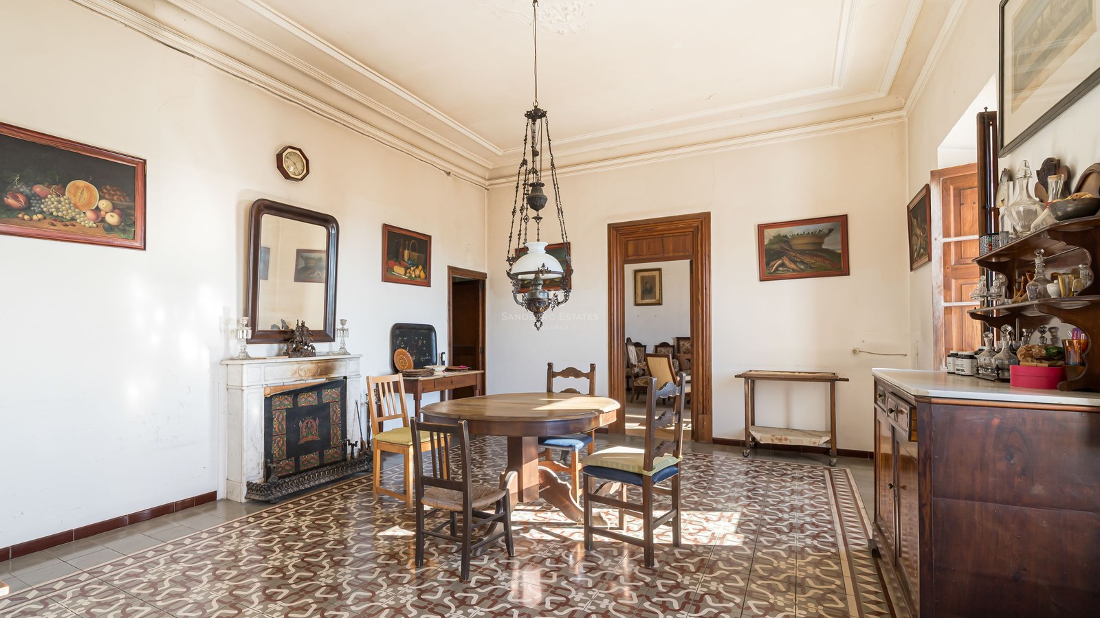 Comedor antiguo con mesa de madera, suelo de baldosas decoradas, chimenea de mármol y techos altos.