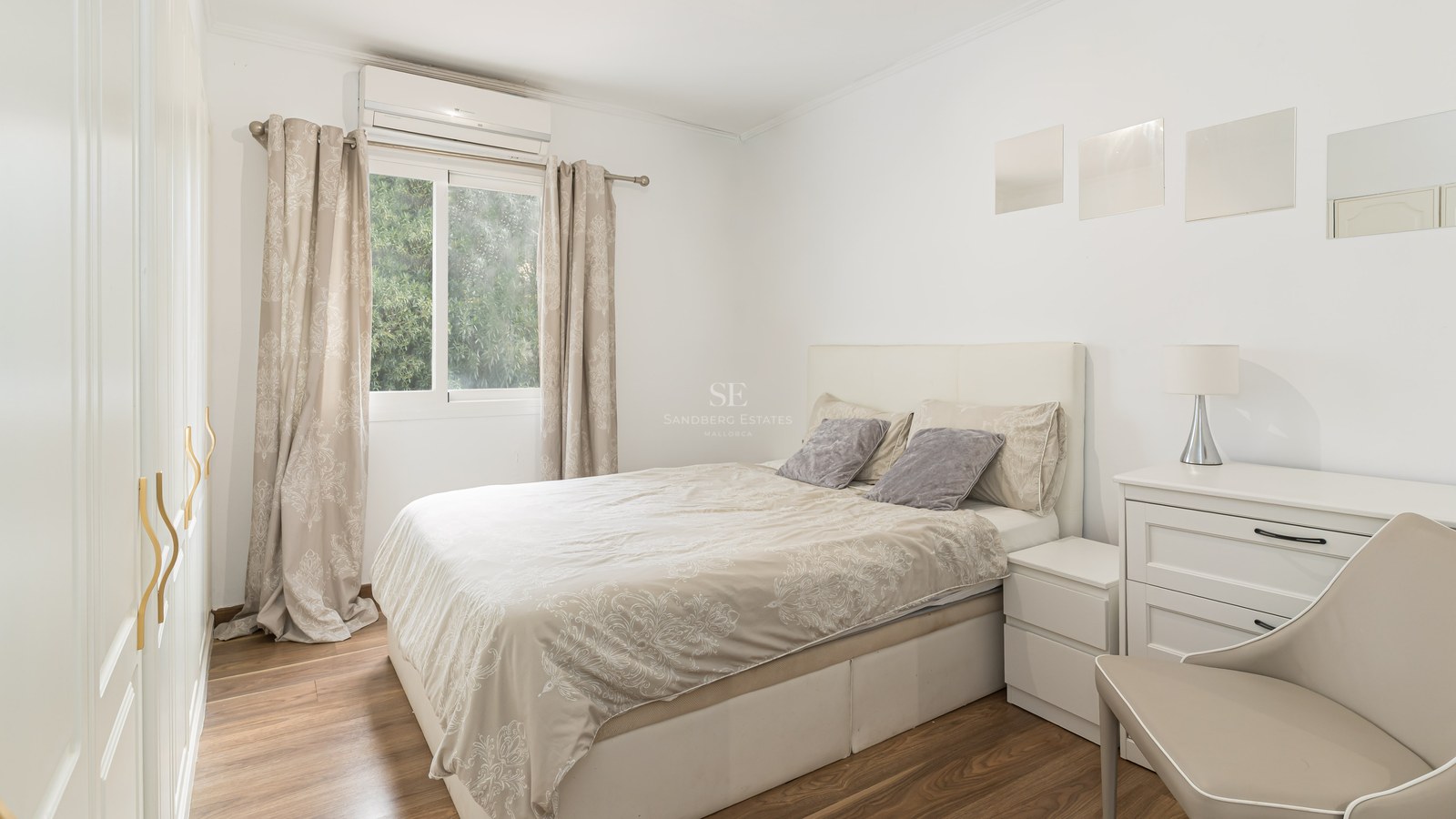 Bright bedroom with a double bed, white furniture, wooden floors, and a window showing green trees.