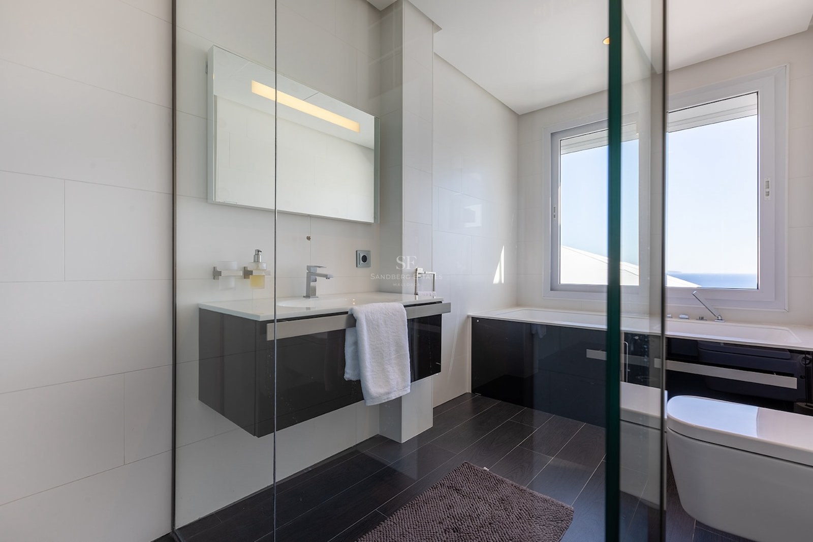 Salle de bain moderne avec murs carrelés blancs, sol sombre, vanité suspendue et baignoire sous une grande fenêtre.
