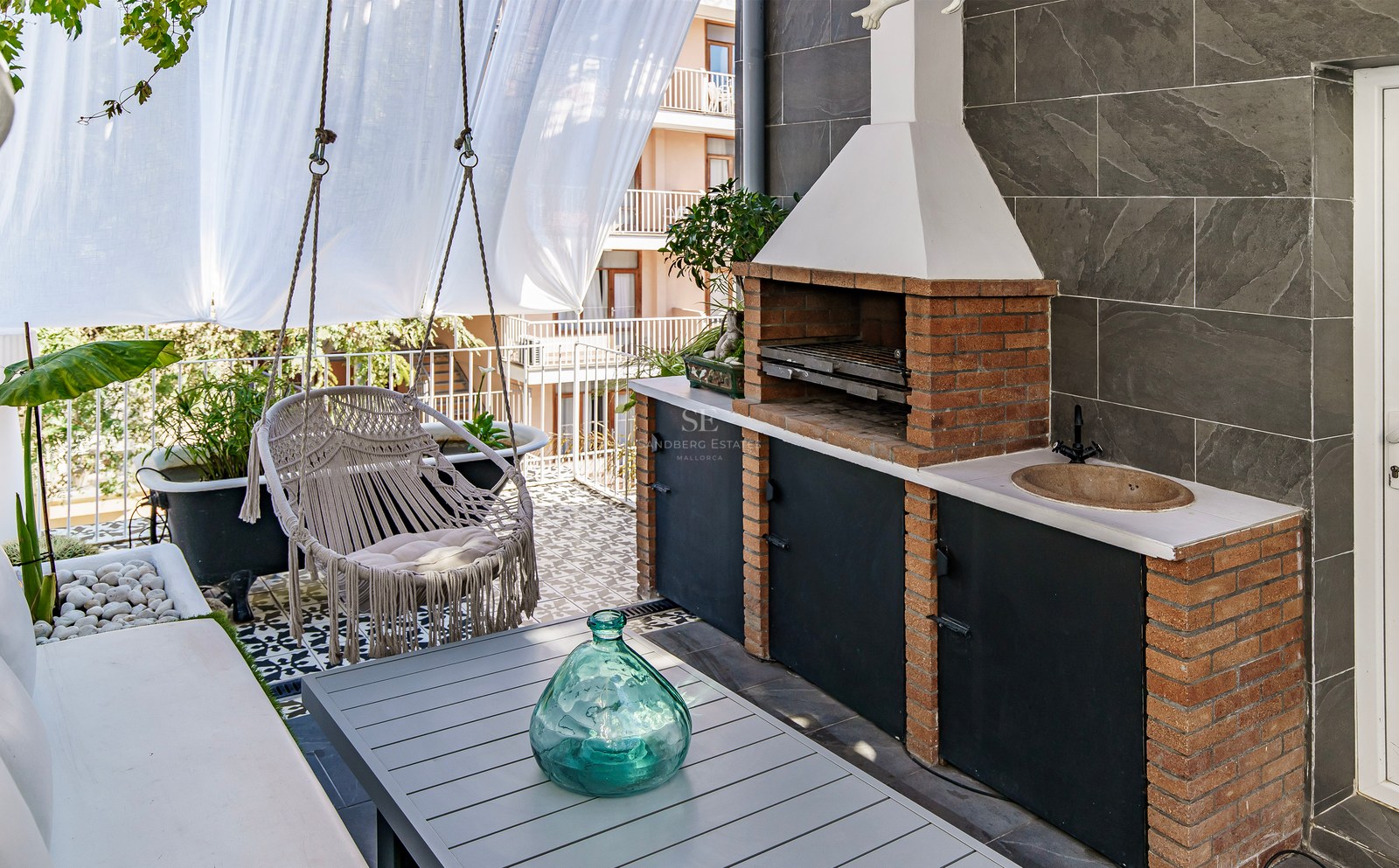 Terrasse moderne avec barbecue en briques, fauteuil suspendu en macramé, table et carrelage décoratif.