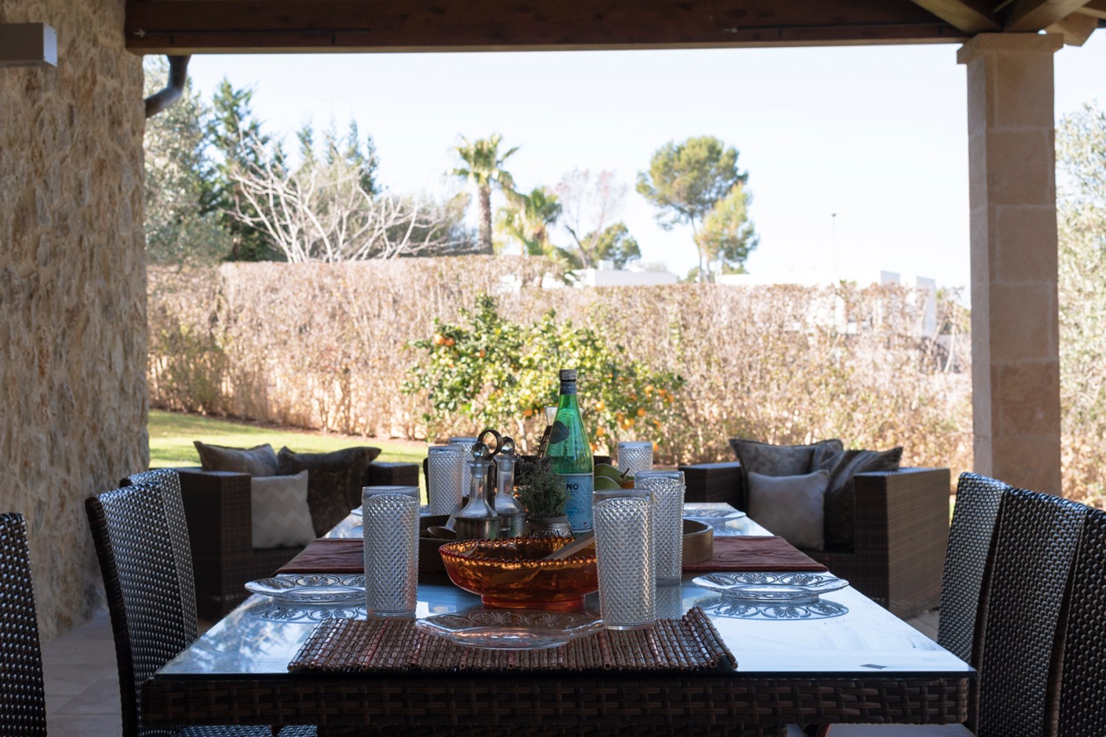 Terrasse méditerranéenne couverte avec salle à manger en osier, murs en pierre naturelle et vue sur un jardin verdoyant.