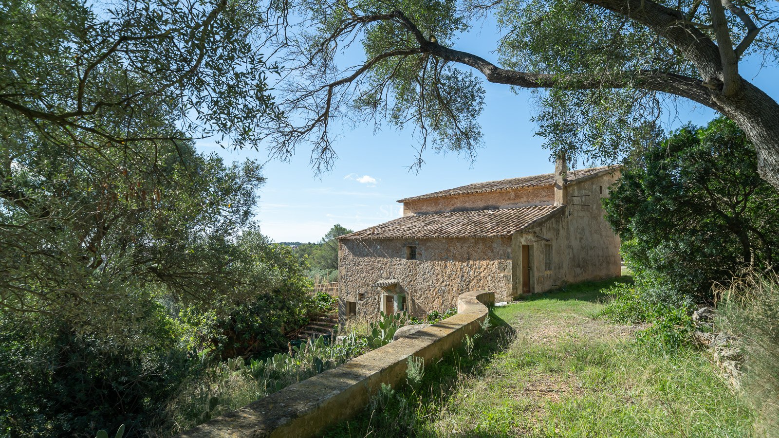 Una casa histórica de piedra de dos plantas con tejado de terracota rodeada de árboles bajo un cielo azul.