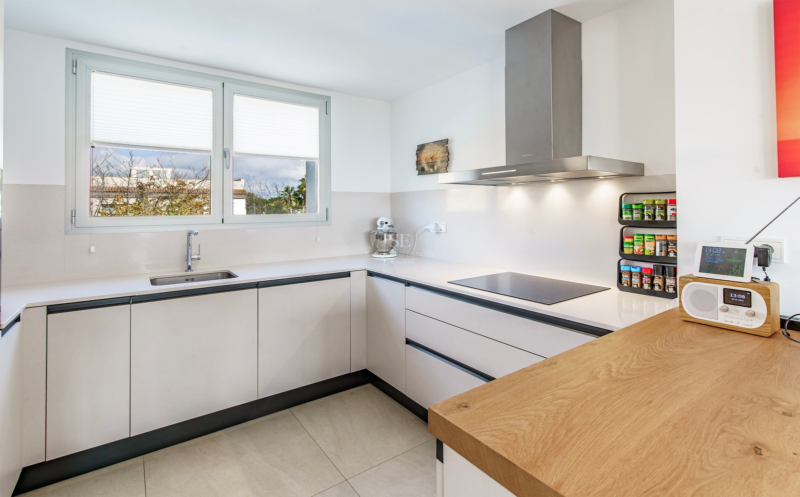 Cuisine blanche moderne en L avec plan de travail en bois, hotte en inox et grande fenêtre.