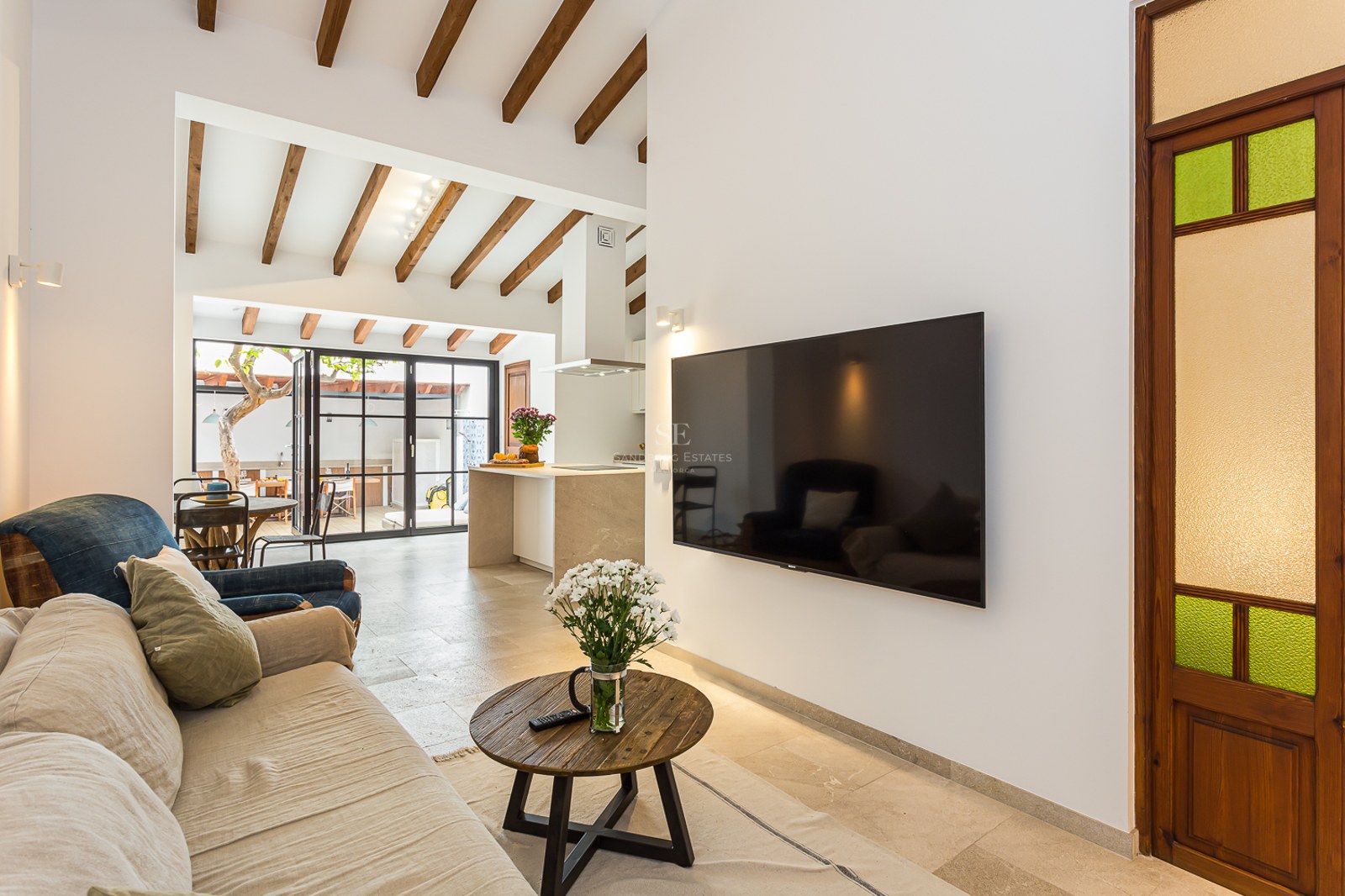 Salón moderno con sofá beige, vigas de madera en el techo, televisor de pantalla plana y vista a la cocina abierta.