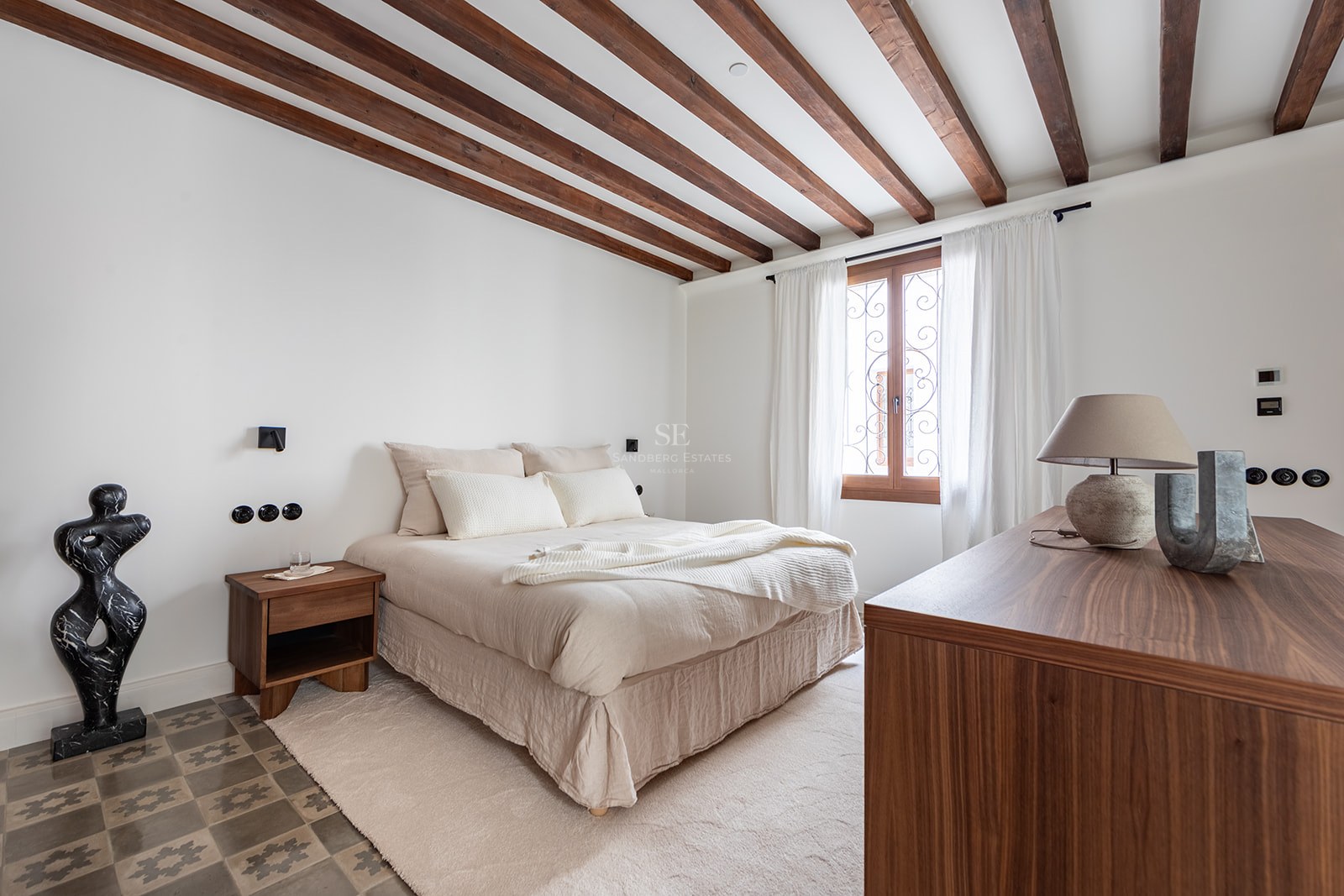 Bedroom featuring exposed dark wood ceiling beams, patterned floor tiles, and a window with sheer white curtains.