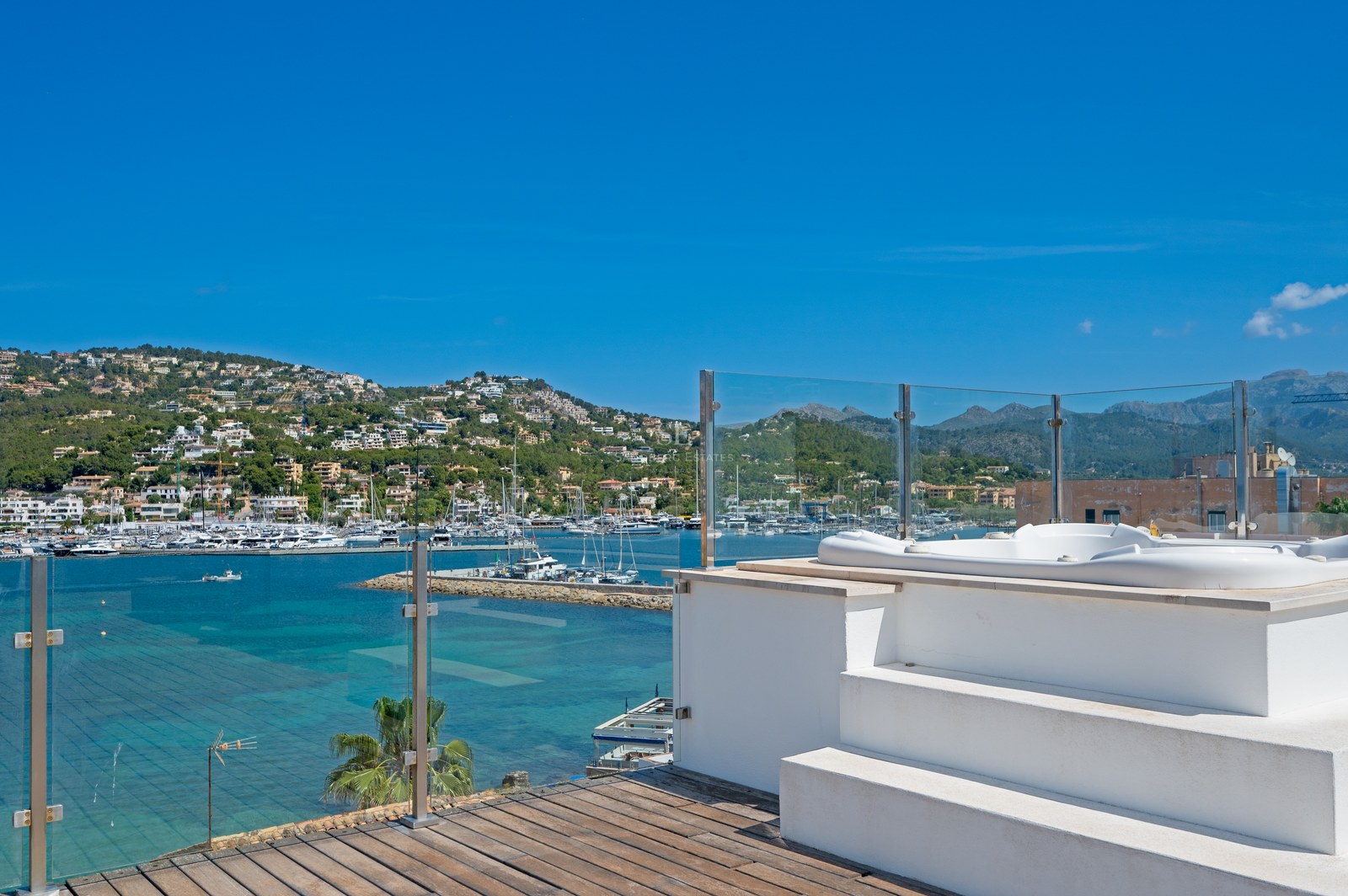 Rooftop terrace featuring a white jacuzzi, glass railings, and panoramic views of a marina and mountains.